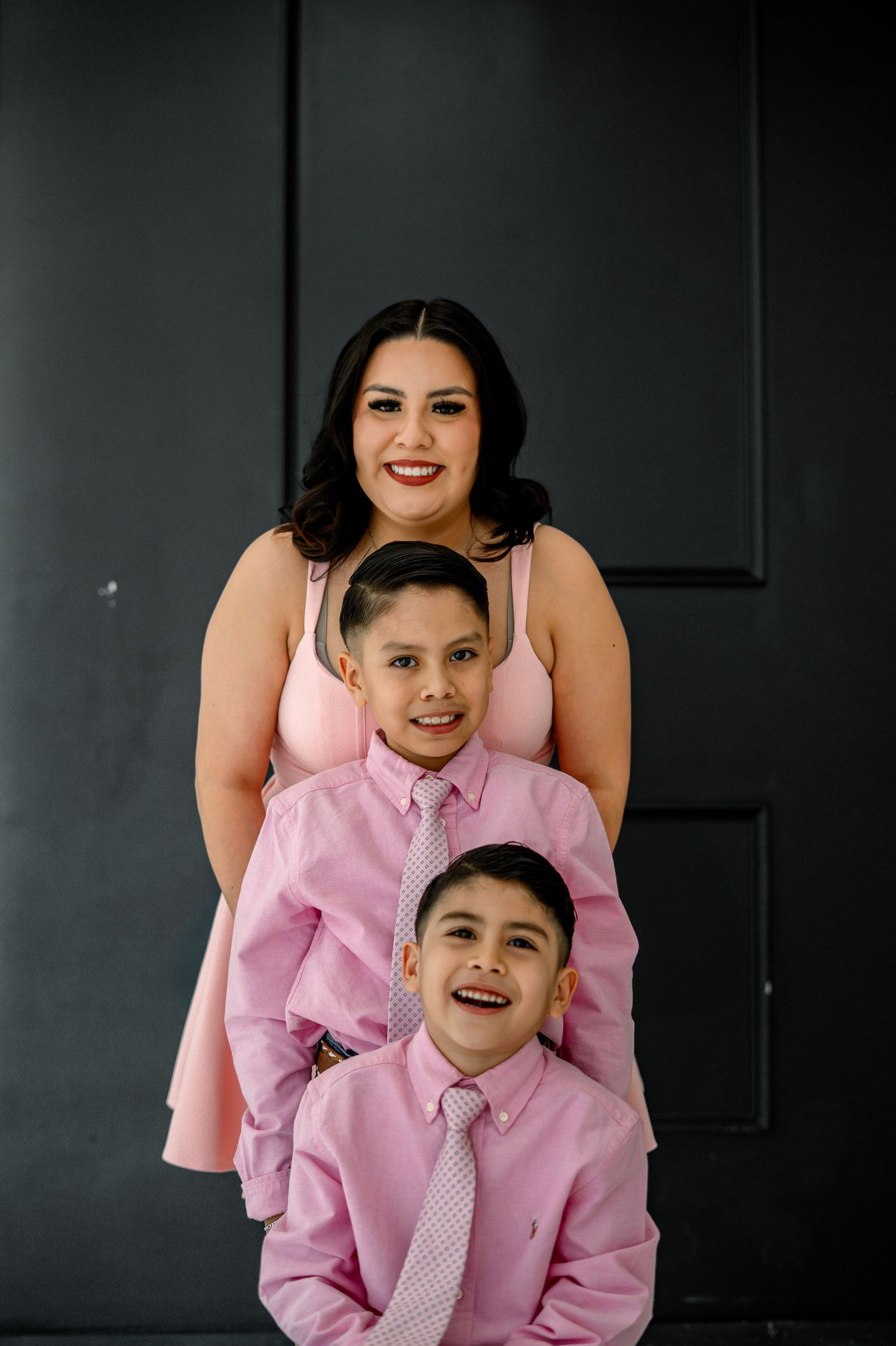 A woman in a pink dress smiles with two young boys in pink shirts and ties in a studio portrait.