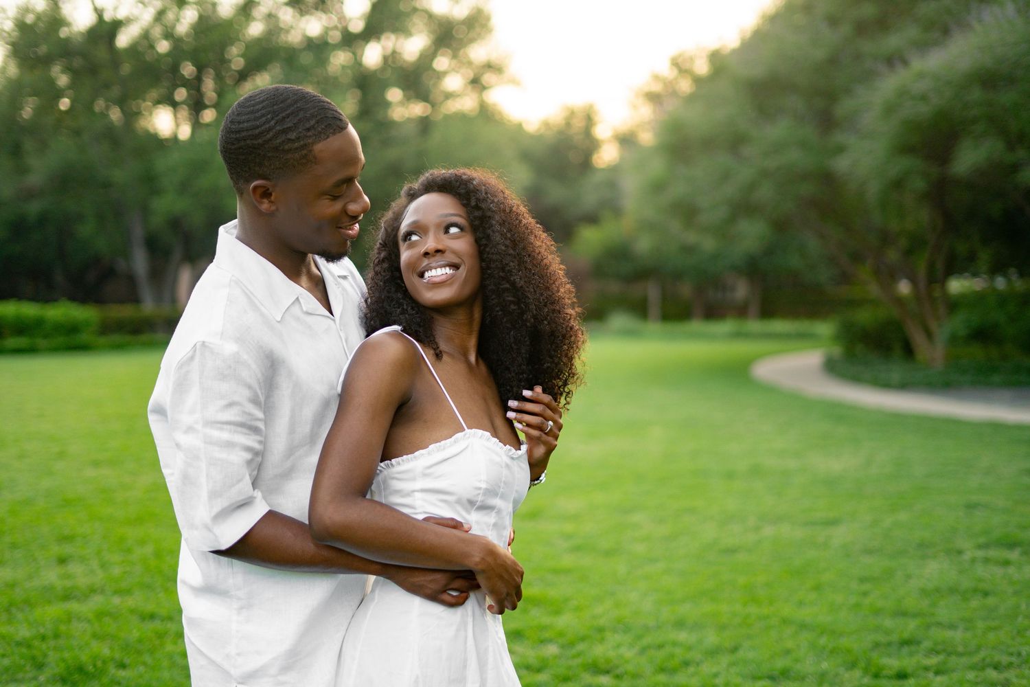 Joyful Black couple in white outfits holding each other close on a green lawn at dusk.