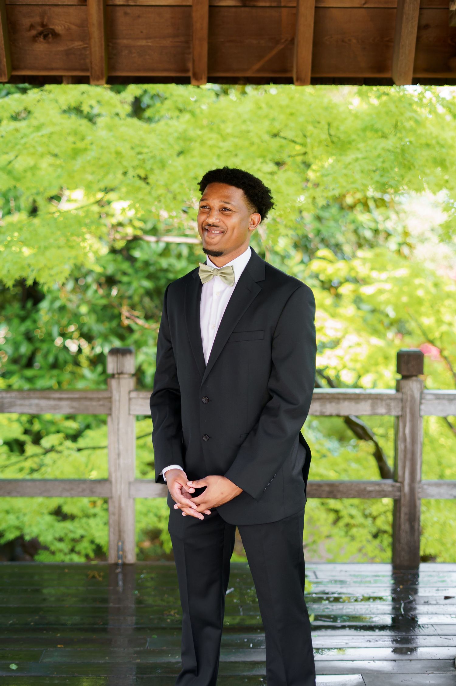 Handsome groom in black tuxedo with gold bow tie smiling under a wooden pavilion surrounded by lush greenery.