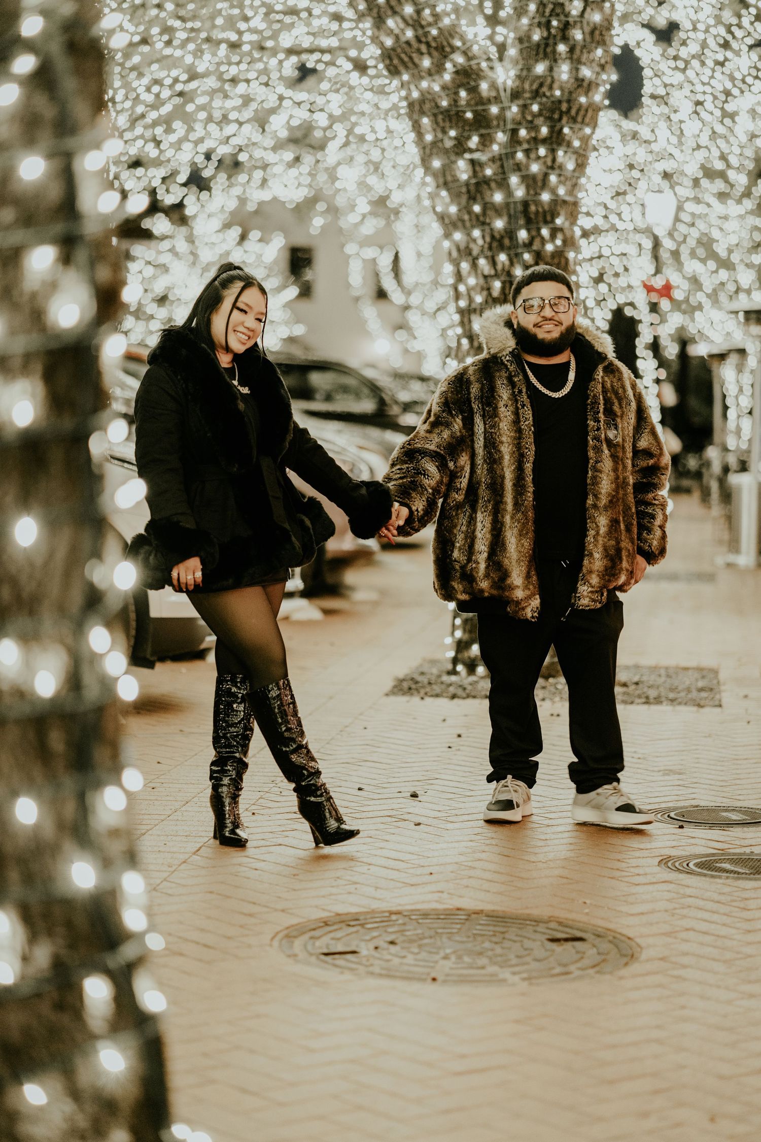 Stylish couple holding hands while walking along a festive brick pathway lined with twinkling lights.