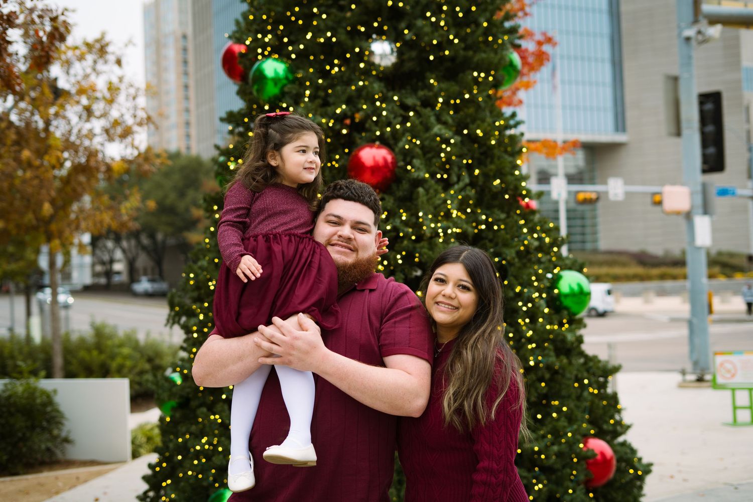 Family of three in matching burgundy outfits poses in front of a large outdoor Christmas tree.