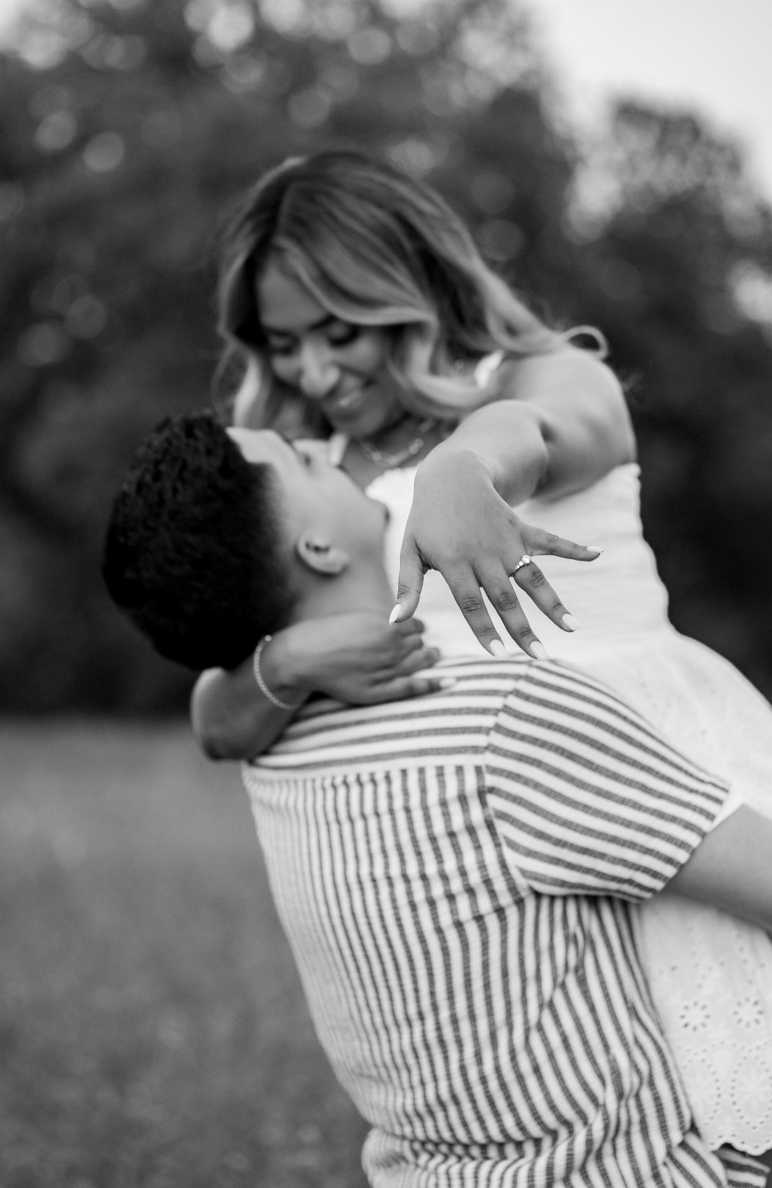 Black and white photo of a man lifting a smiling woman wearing an engagement ring outdoors.