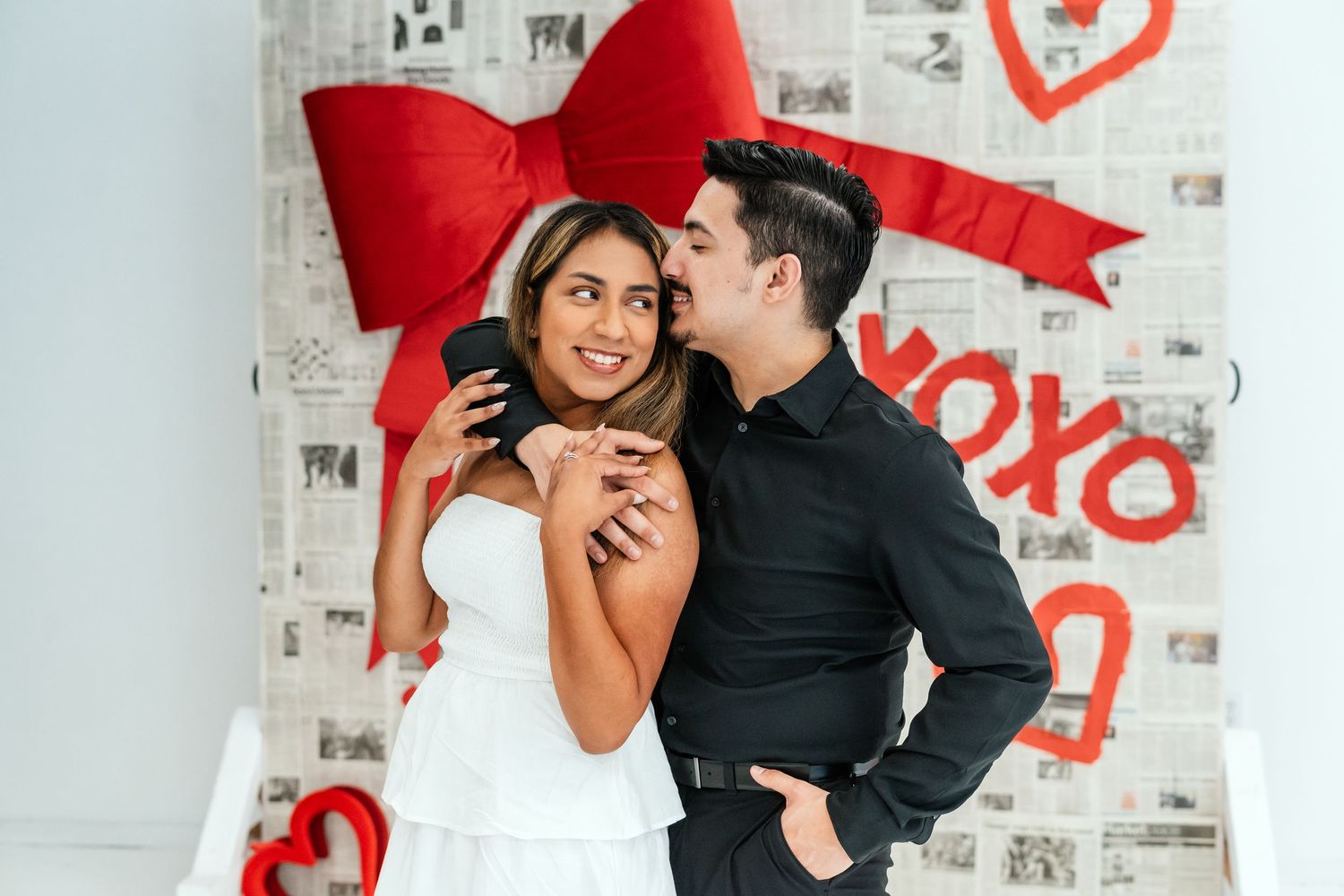 Man embracing woman from behind in front of Valentine's Day XOXO newspaper backdrop with red hearts.