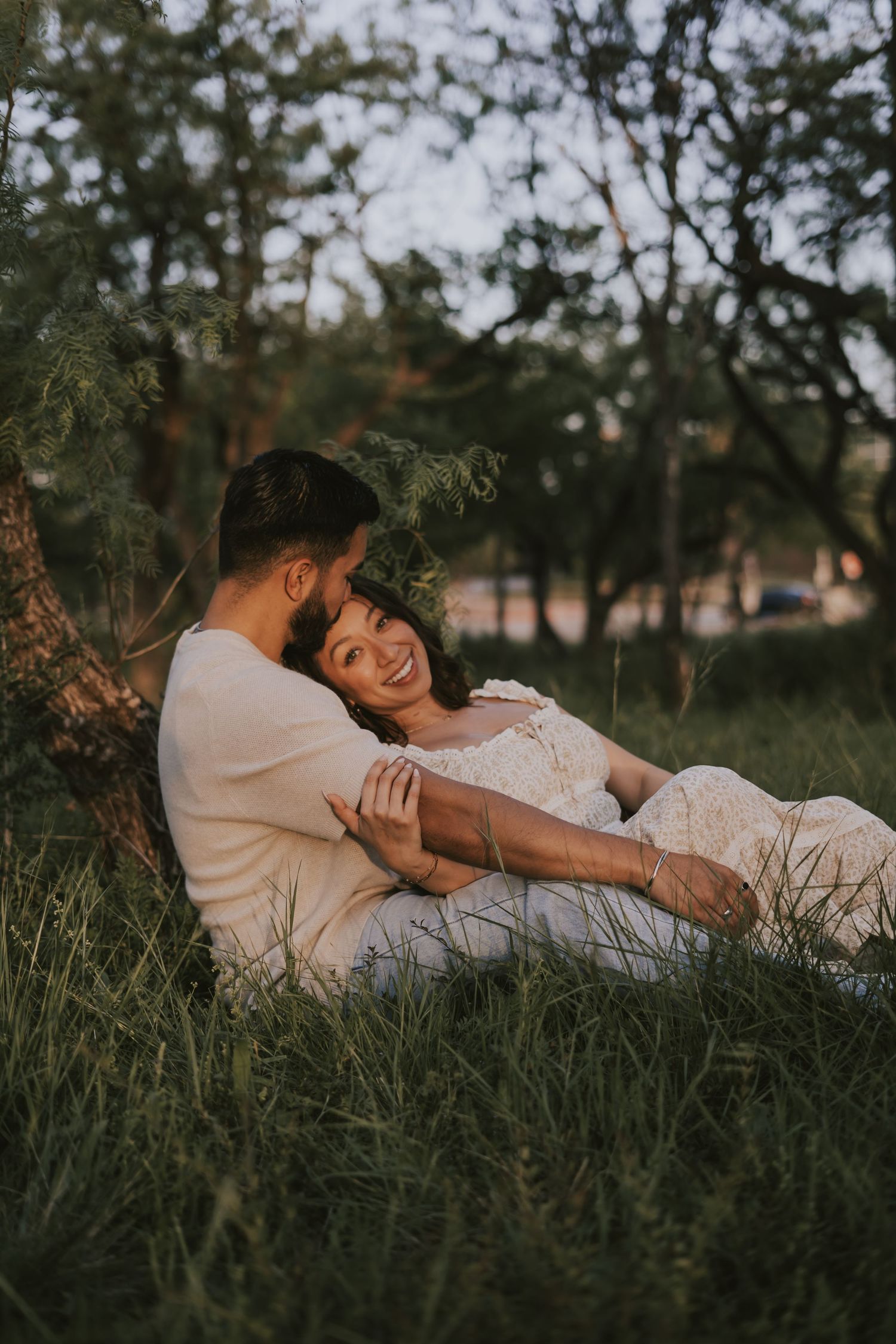 A couple sits together in lush green grass leaning against a tree, sharing a tender romantic moment outdoors.