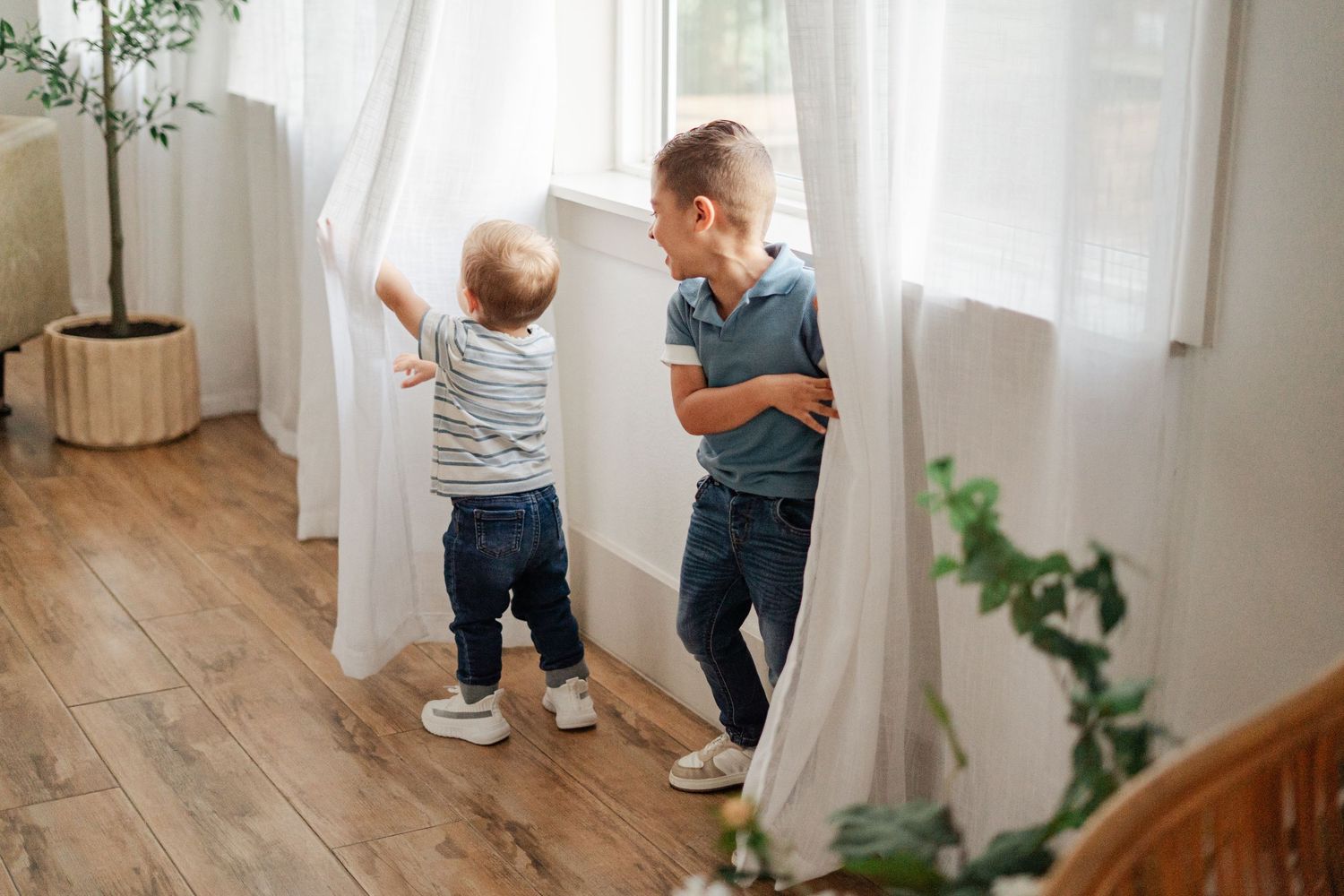 Two young boys playing near white curtains by a sunny window on a hardwood floor indoors.