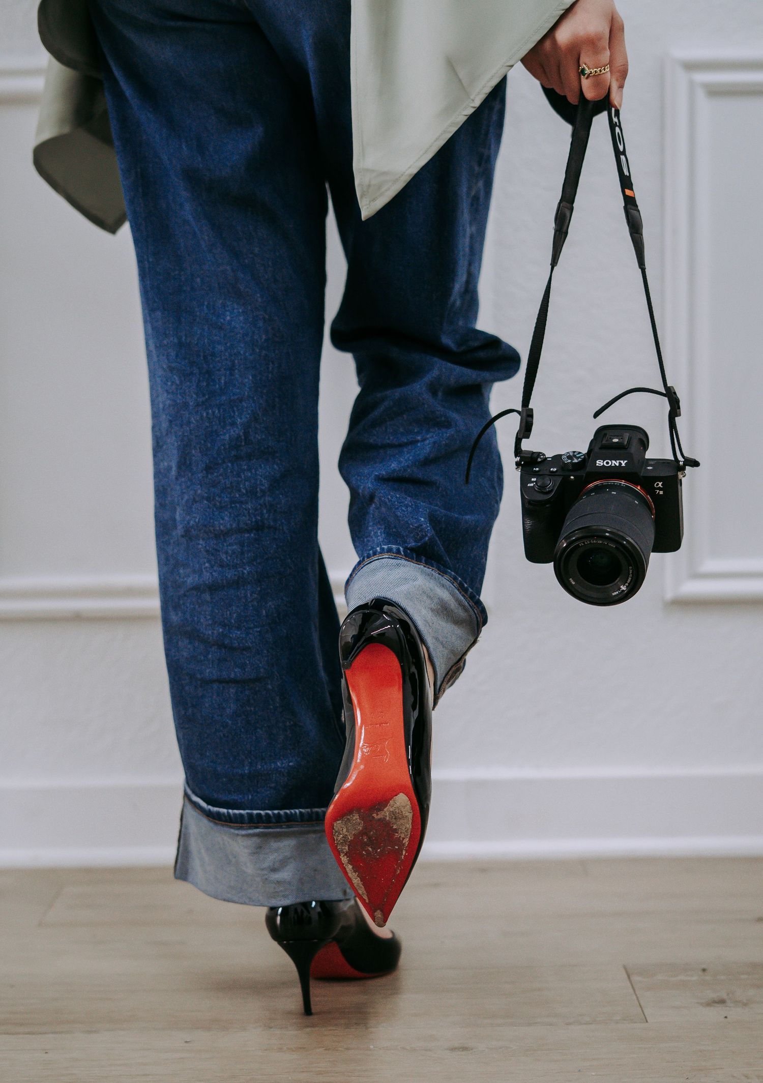 Person walking in black Louboutin heels and cuffed jeans, carrying a Sony mirrorless camera by its strap.