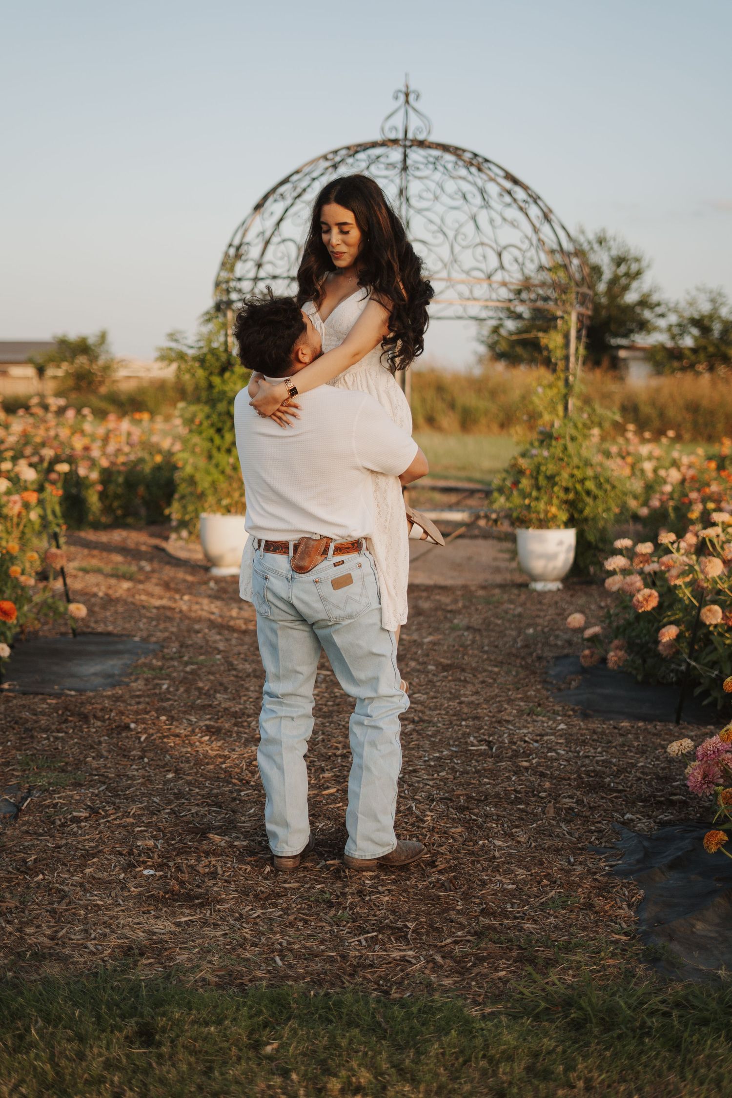 Couple embracing in a romantic rose garden with an ornate metal arbor backdrop at golden hour.