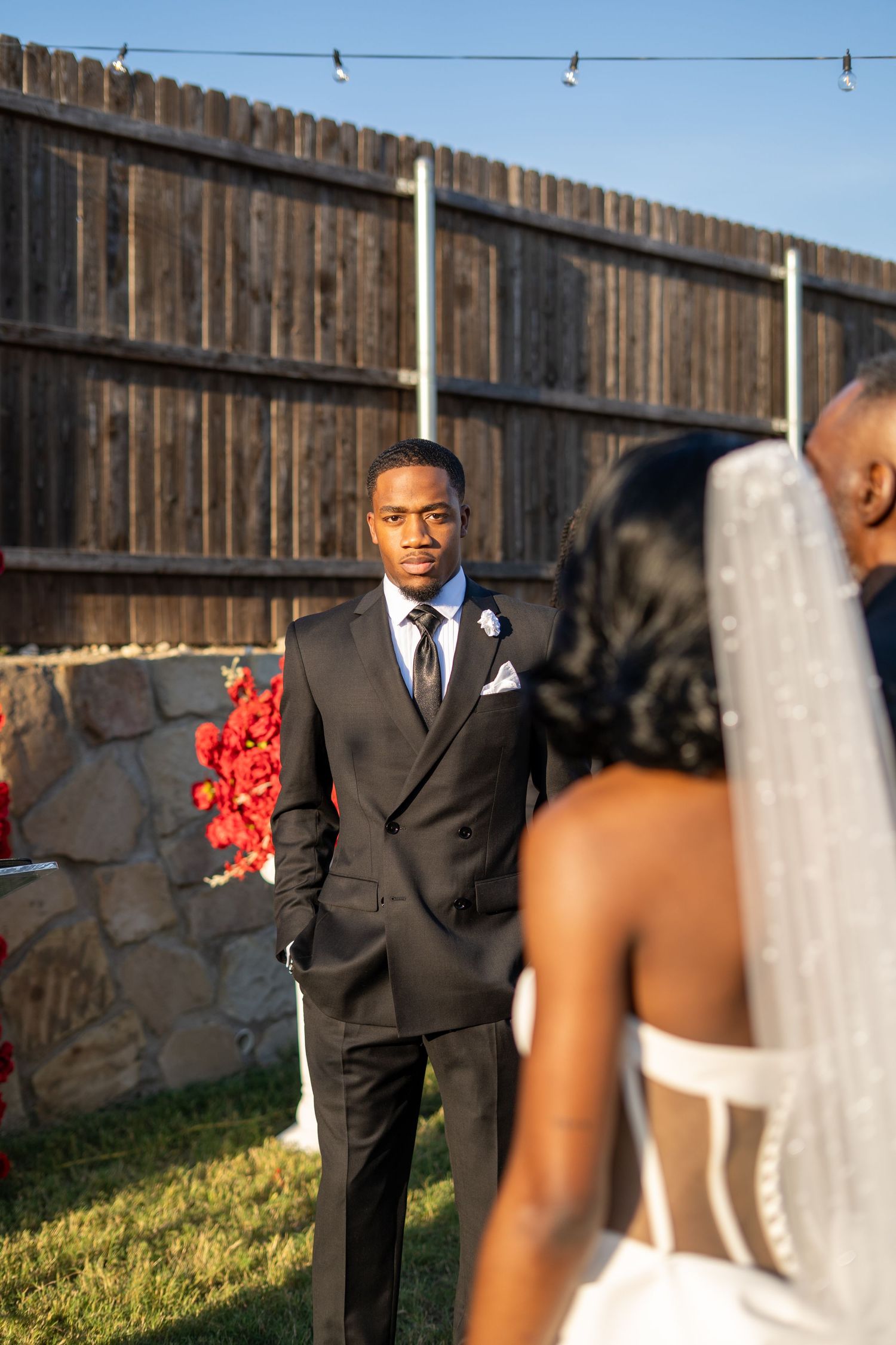 Groom in black suit watches intently as bride in white gown and veil approaches outdoor ceremony.