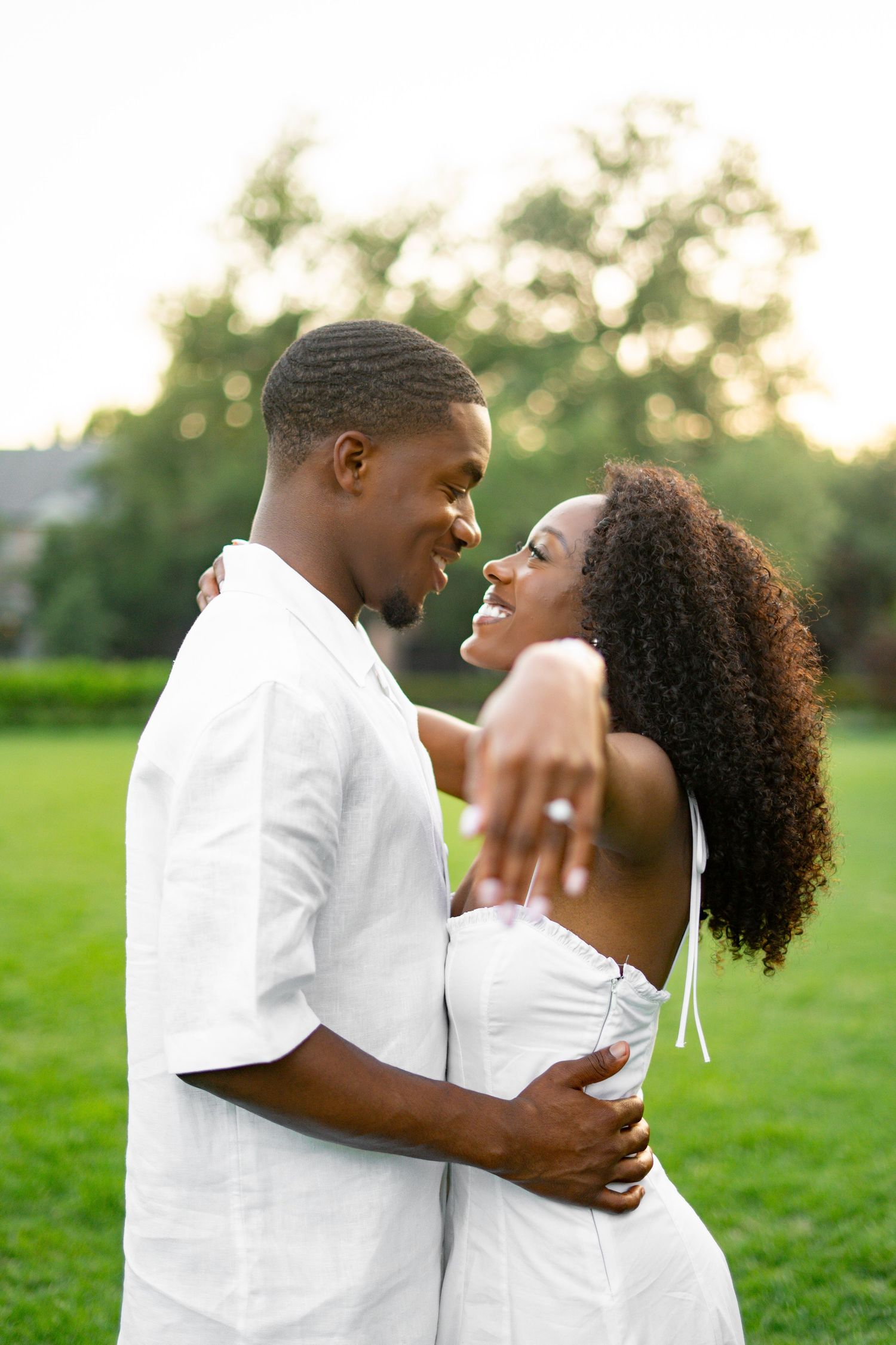 Happy Black couple in white outfits facing each other, woman showing engagement ring on green grass.