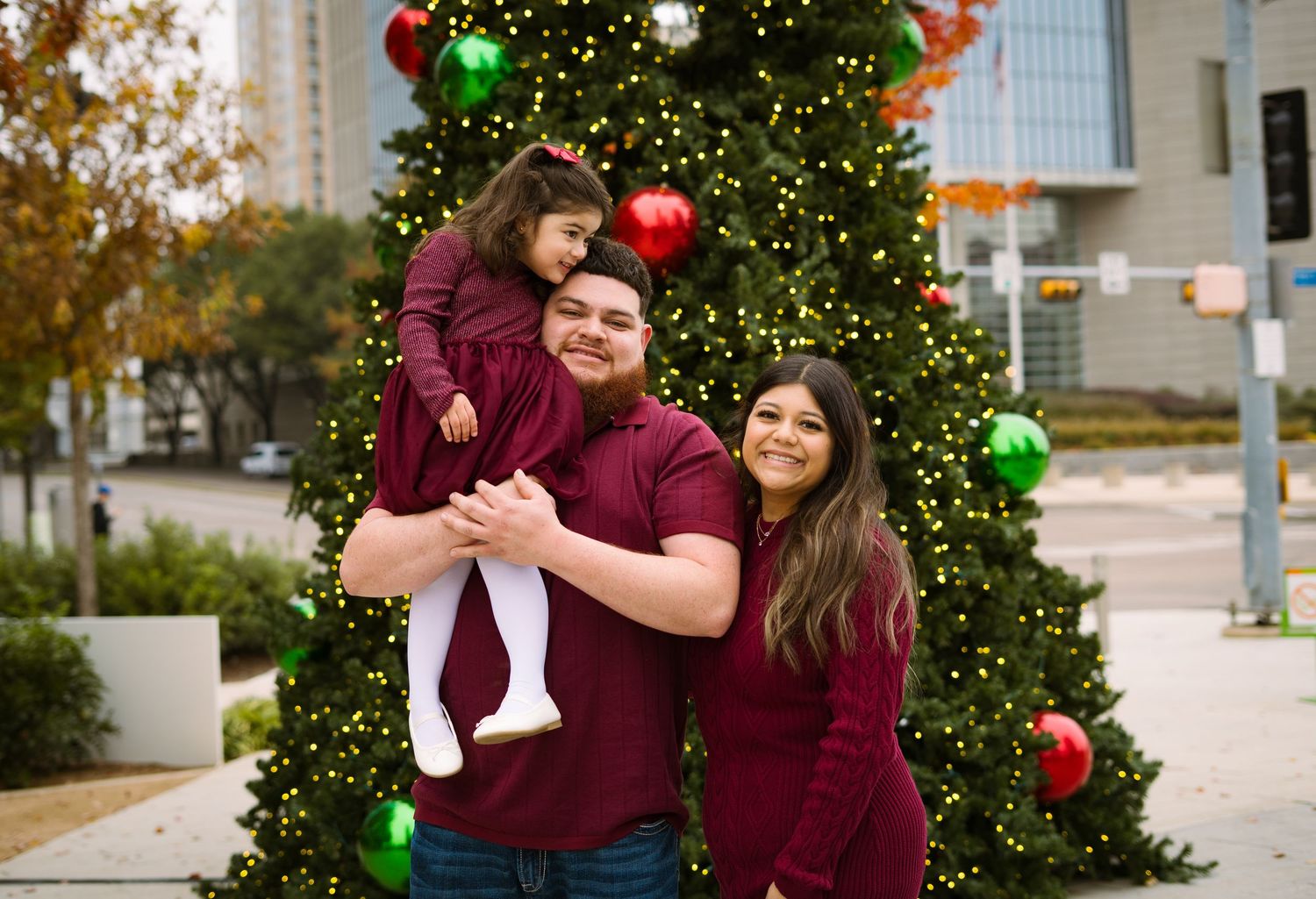 Smiling family of three in matching burgundy clothing poses together in front of a Christmas tree.