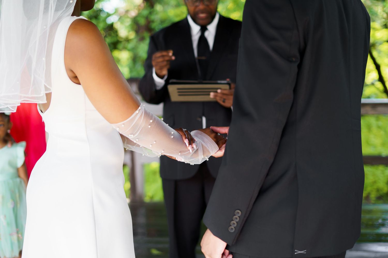 Bride and groom hold hands during wedding ceremony as officiant reads from book under wooden pavilion.
