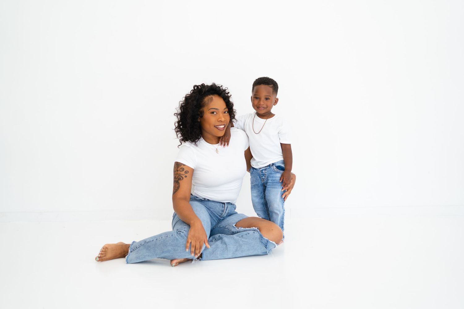 Black mother sitting on floor with her toddler son in matching white tops and jeans in studio.
