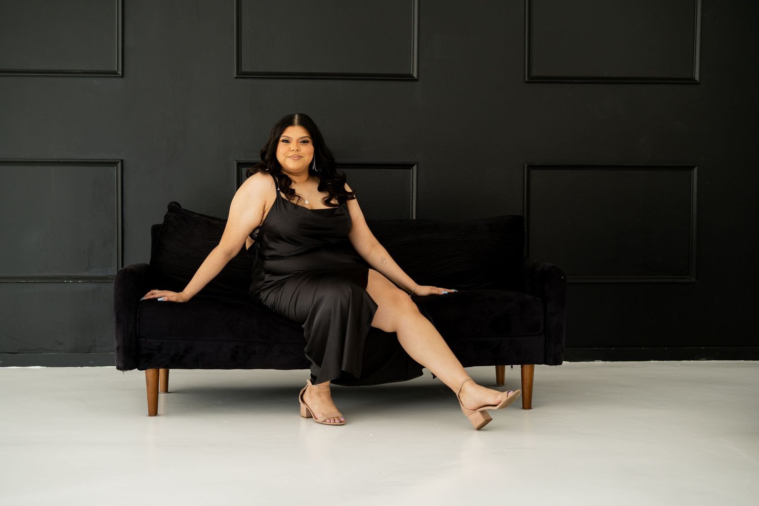 A woman in a black satin dress poses elegantly on a velvet sofa against a dark wall.