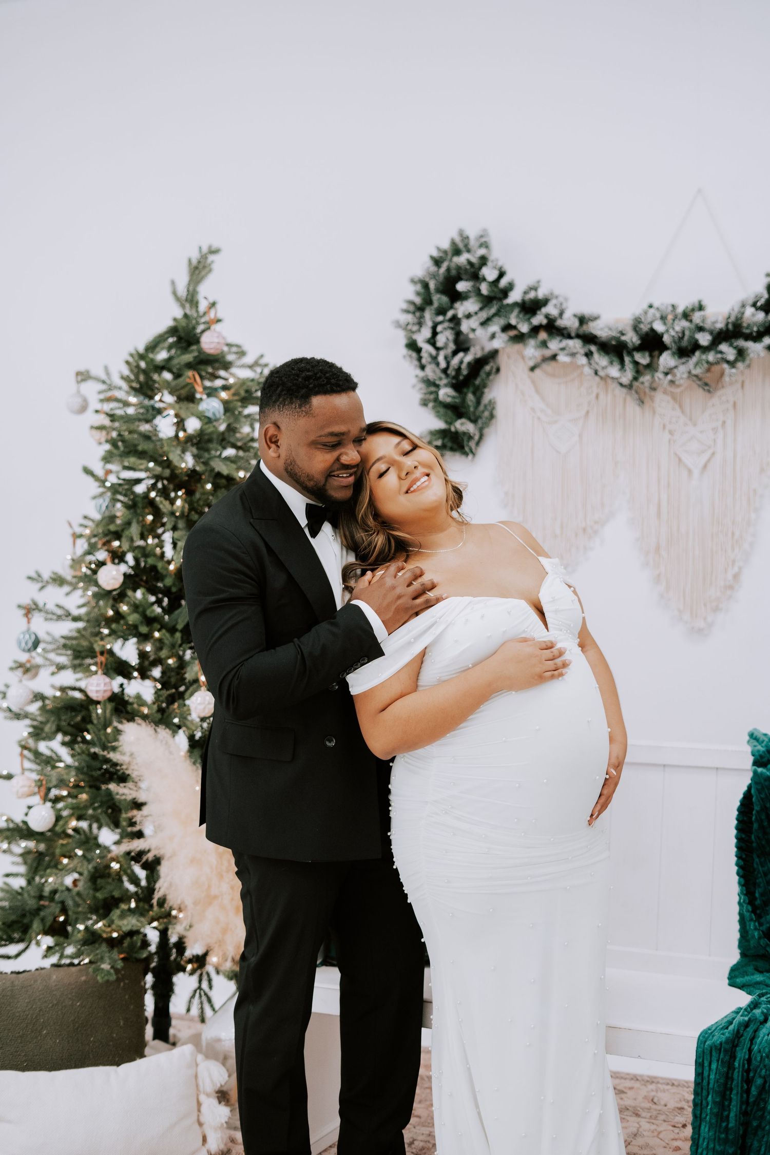 Couple in wedding attire embracing near Christmas tree, pregnant bride in white gown with groom in black tuxedo.