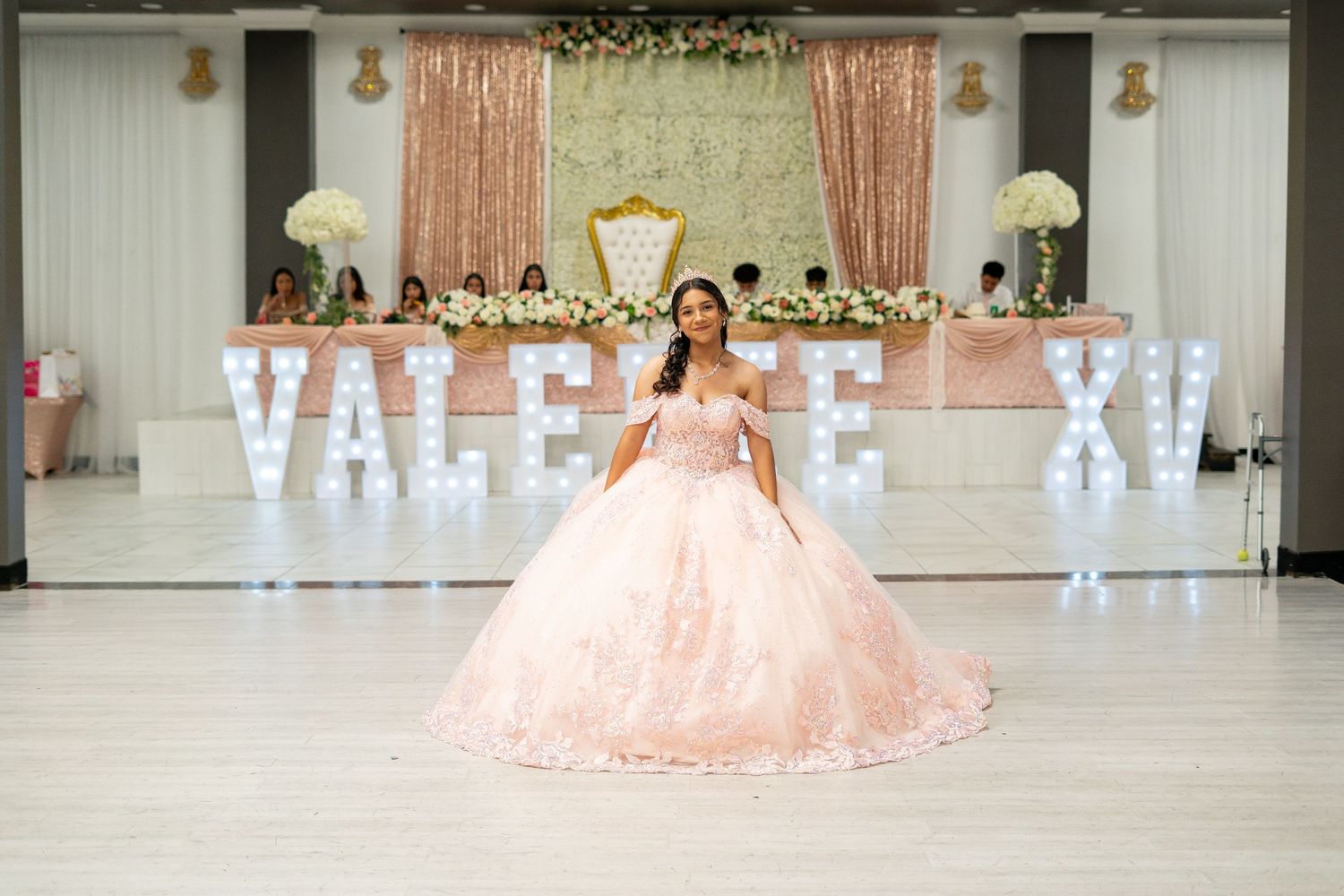 Young woman in pink ball gown poses at her Quinceañera in front of VALERIE XV marquee letters.