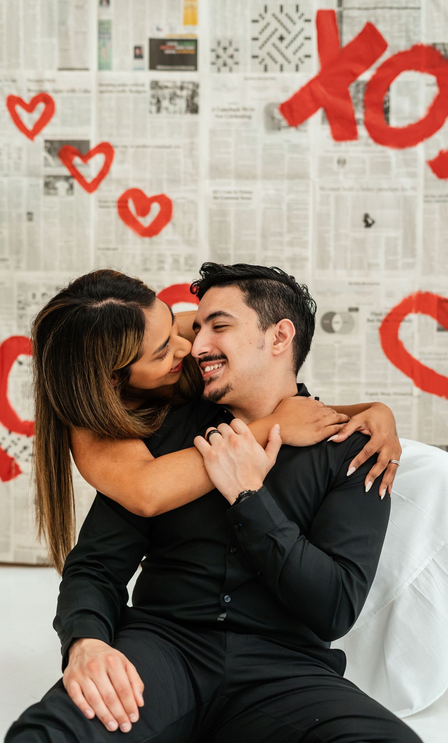 Woman hugging smiling man seated on white couch in front of XOXO Valentine's Day backdrop.