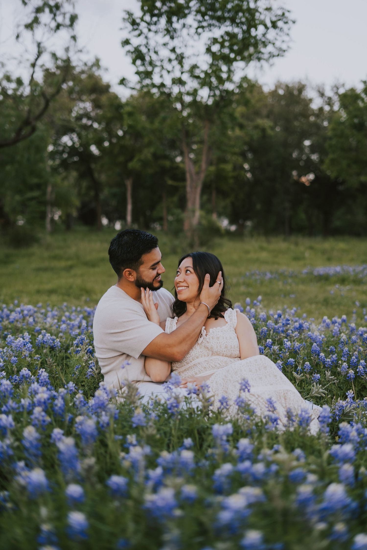 Couple sitting in a field of blue wildflowers, sharing a tender moment surrounded by lush green trees.