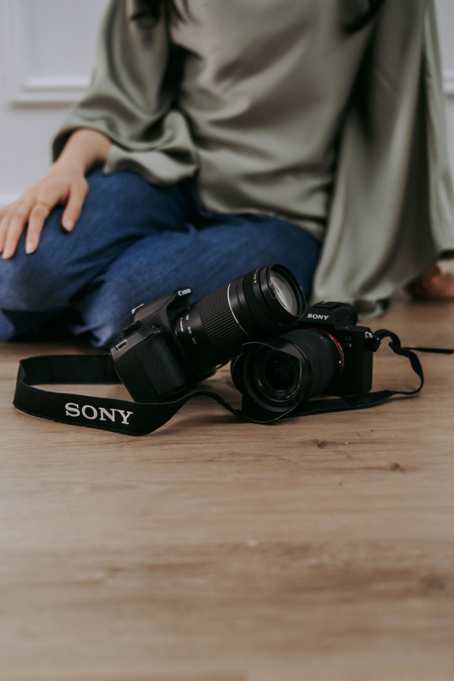 Two Sony cameras with lenses on a wooden floor beside a person sitting in jeans and an olive green top.