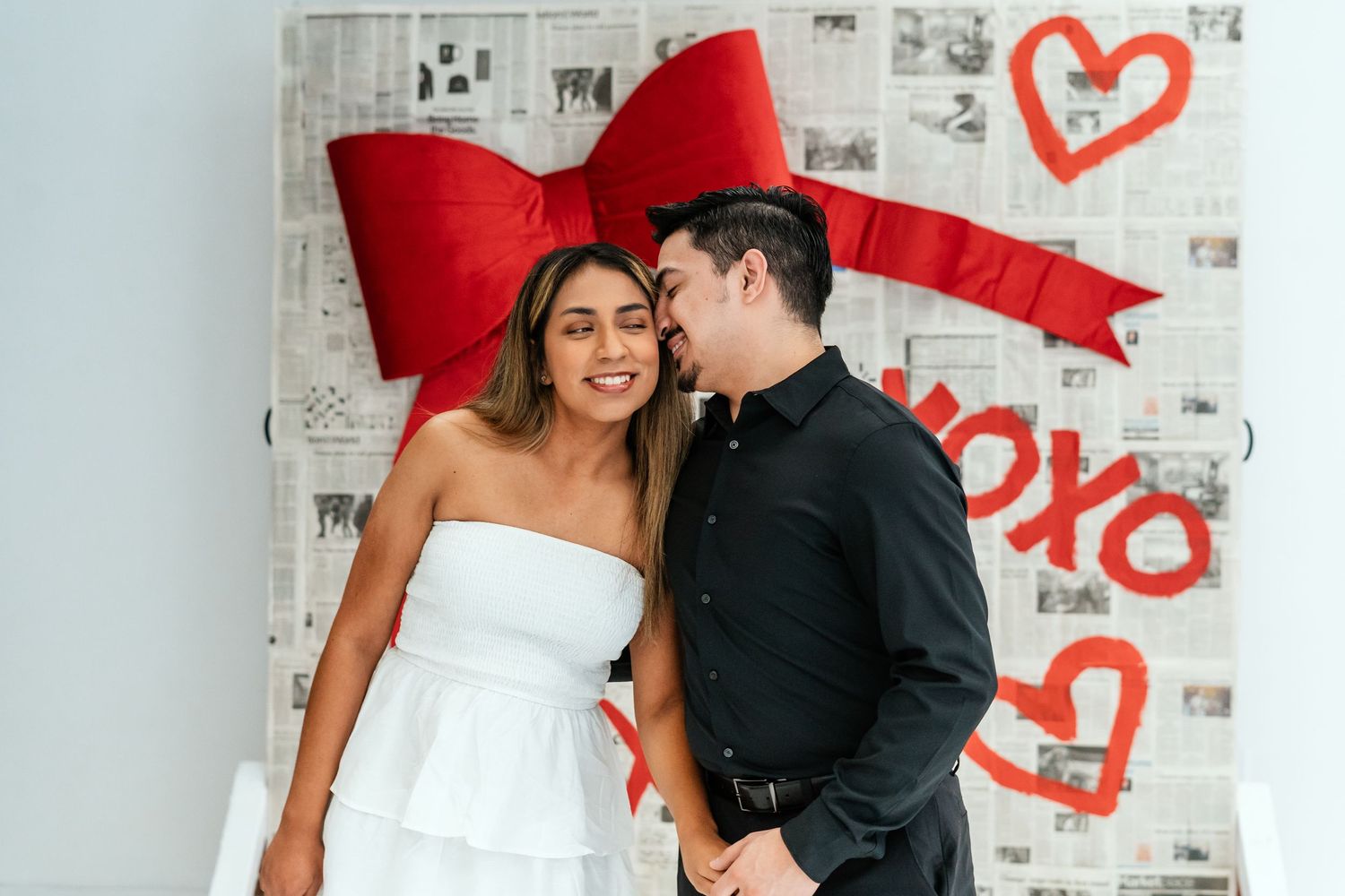 Couple sharing a romantic moment in front of XOXO Valentine's Day backdrop with large red bow.