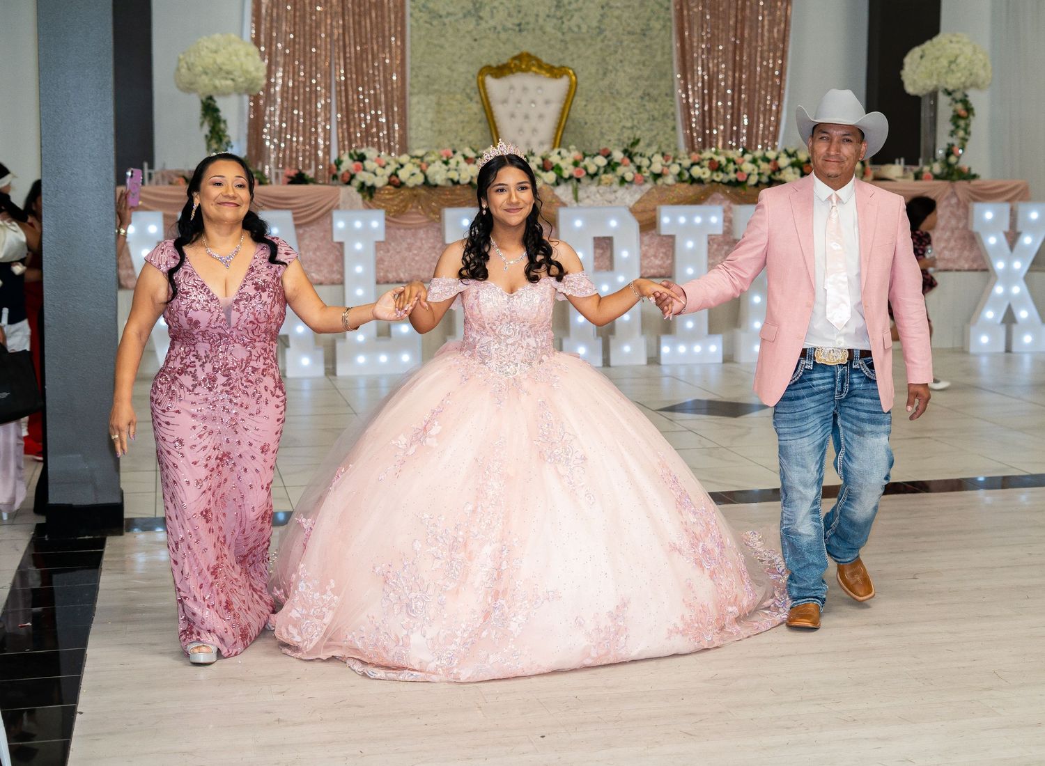 A quinceañera in a pink ball gown walks with her parents during her elegant celebration.