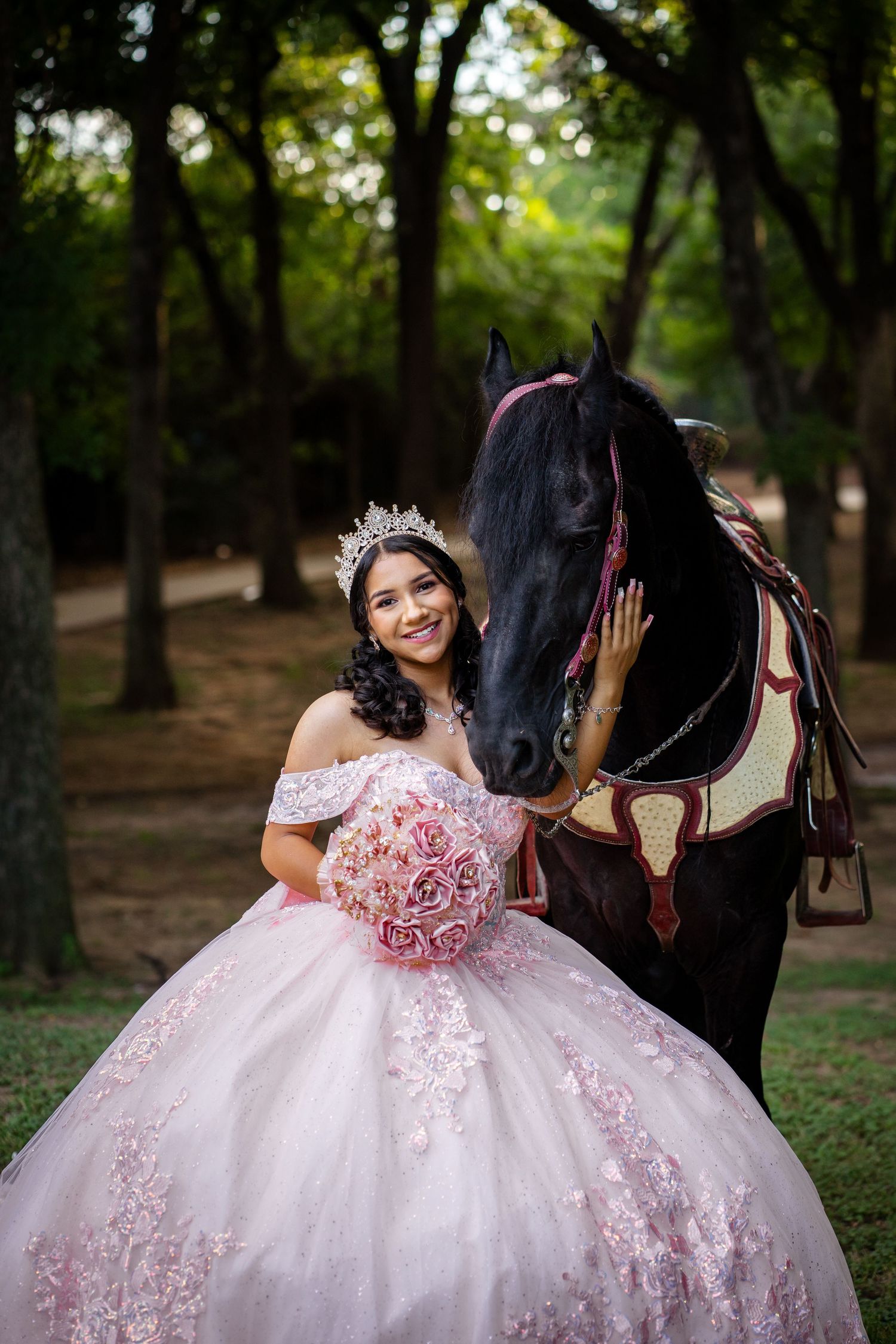 Young woman in pink quinceañera ball gown with tiara and floral bouquet posing beside a black horse outdoors.