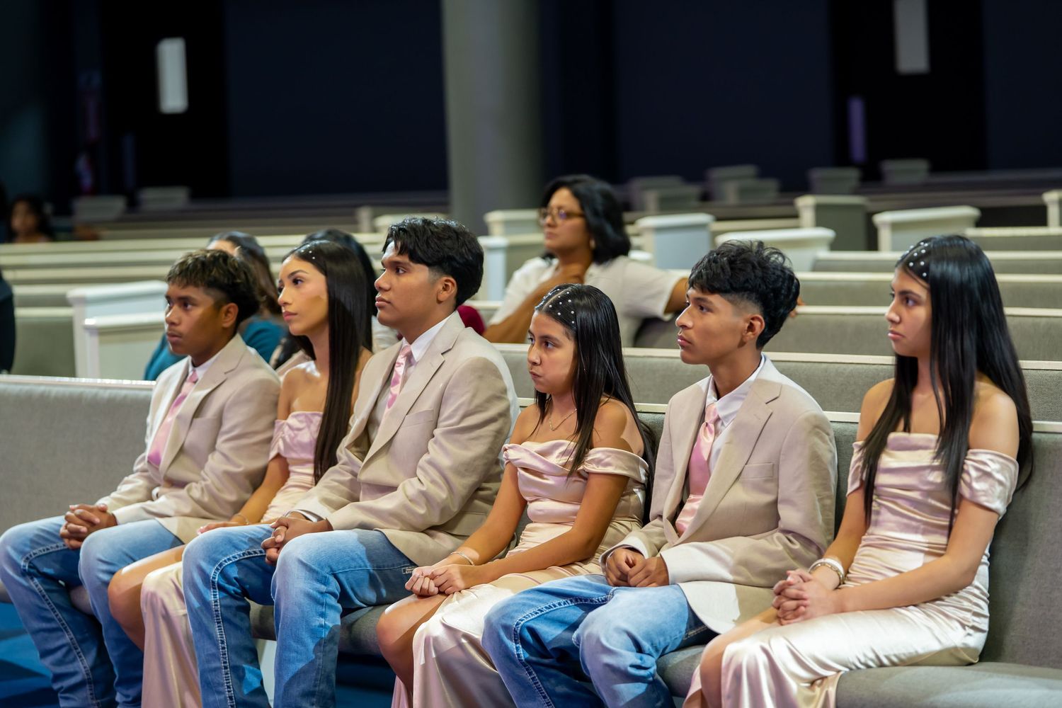 Group of six teenagers in formal attire seated in church pews during a quinceañera ceremony.