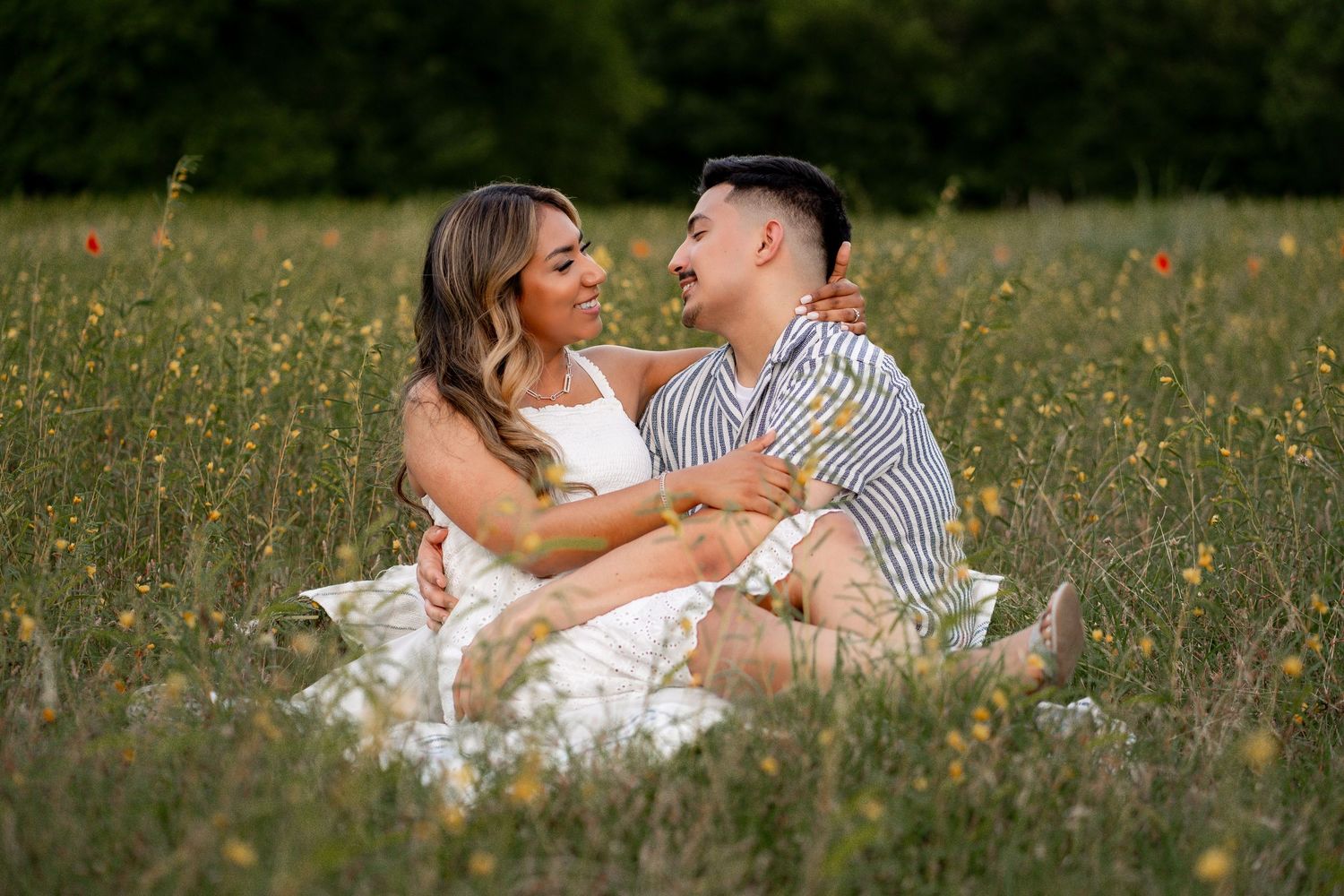 Happy couple sitting together in a wildflower meadow, smiling and embracing during an outdoor engagement session.