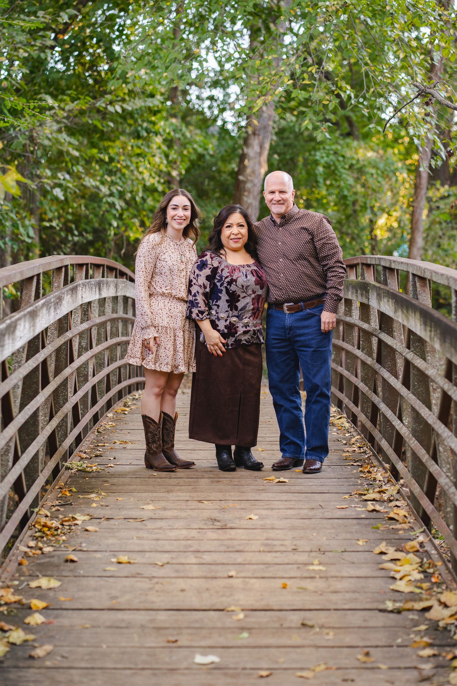 Three family members smiling together on a wooden bridge covered in autumn leaves.