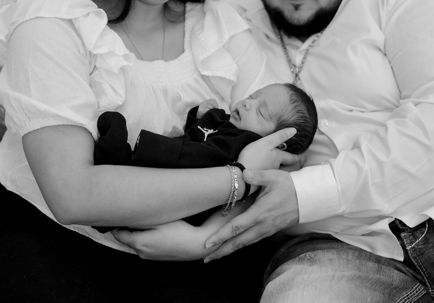 Black and white photo of parents cradling sleeping newborn baby dressed in black outfit.
