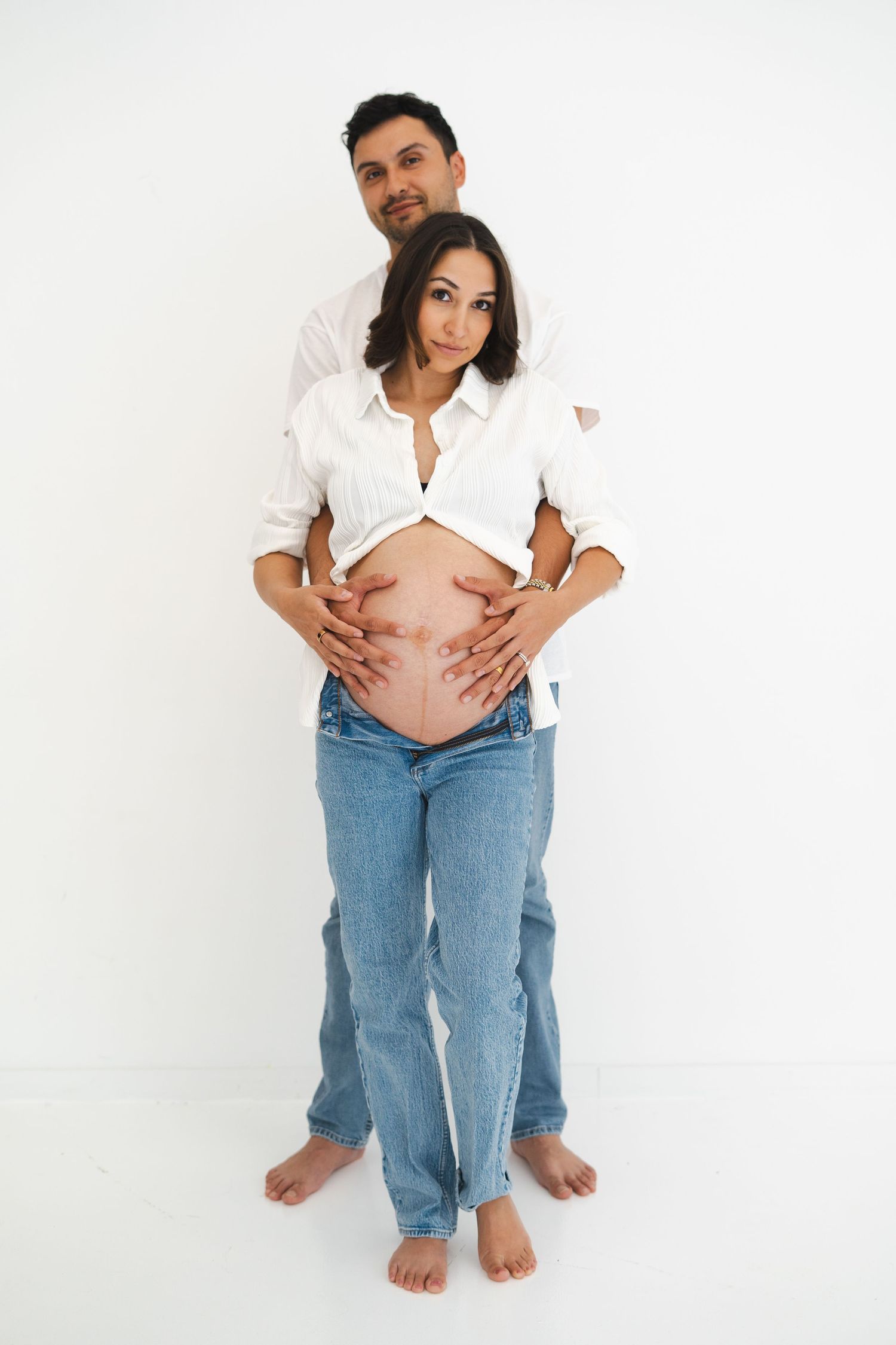 Man embracing pregnant partner from behind, both in white shirts and jeans, studio photo.