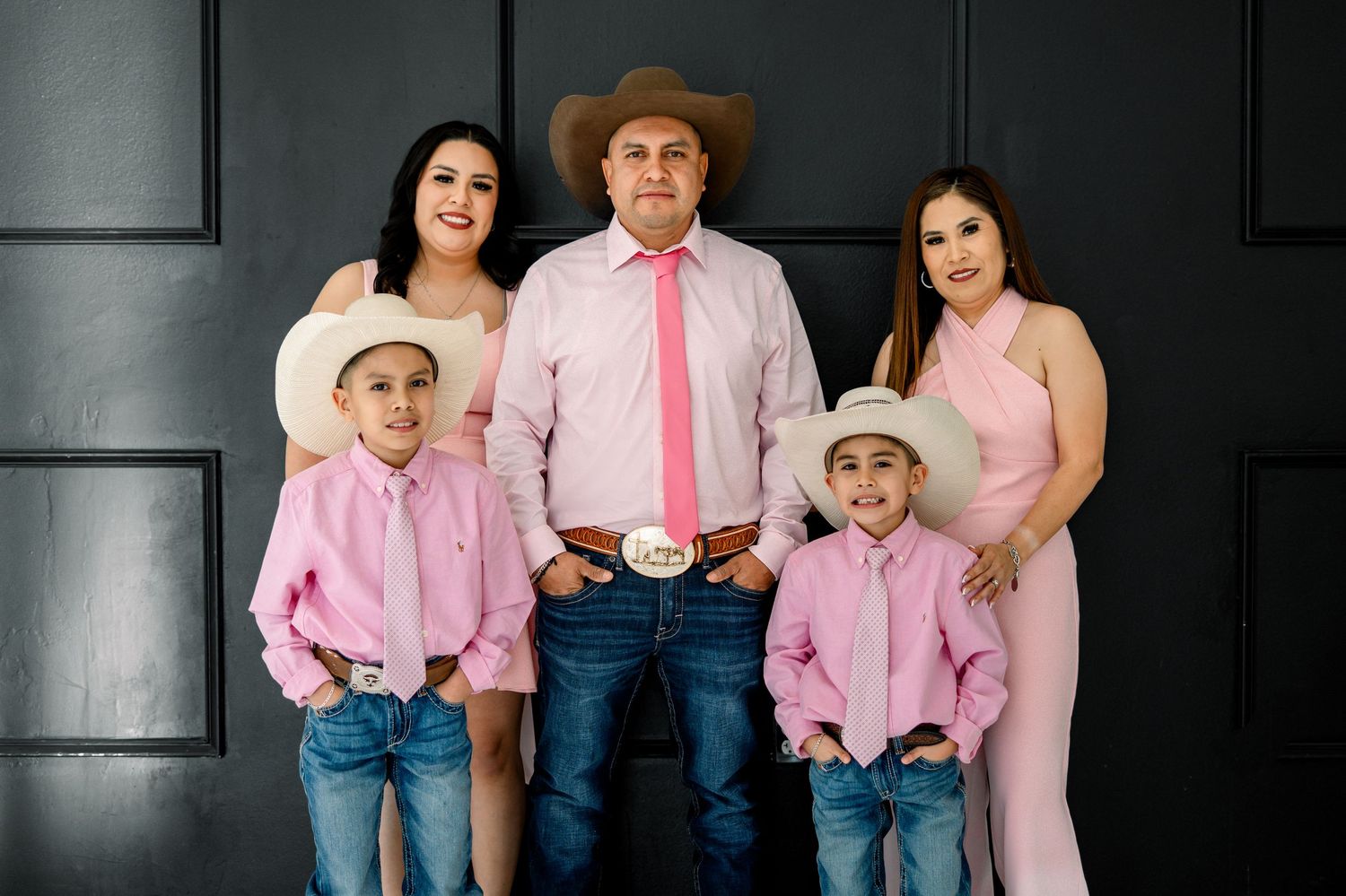 A family of five in coordinated pink outfits with cowboy hats pose together against a dark wall.