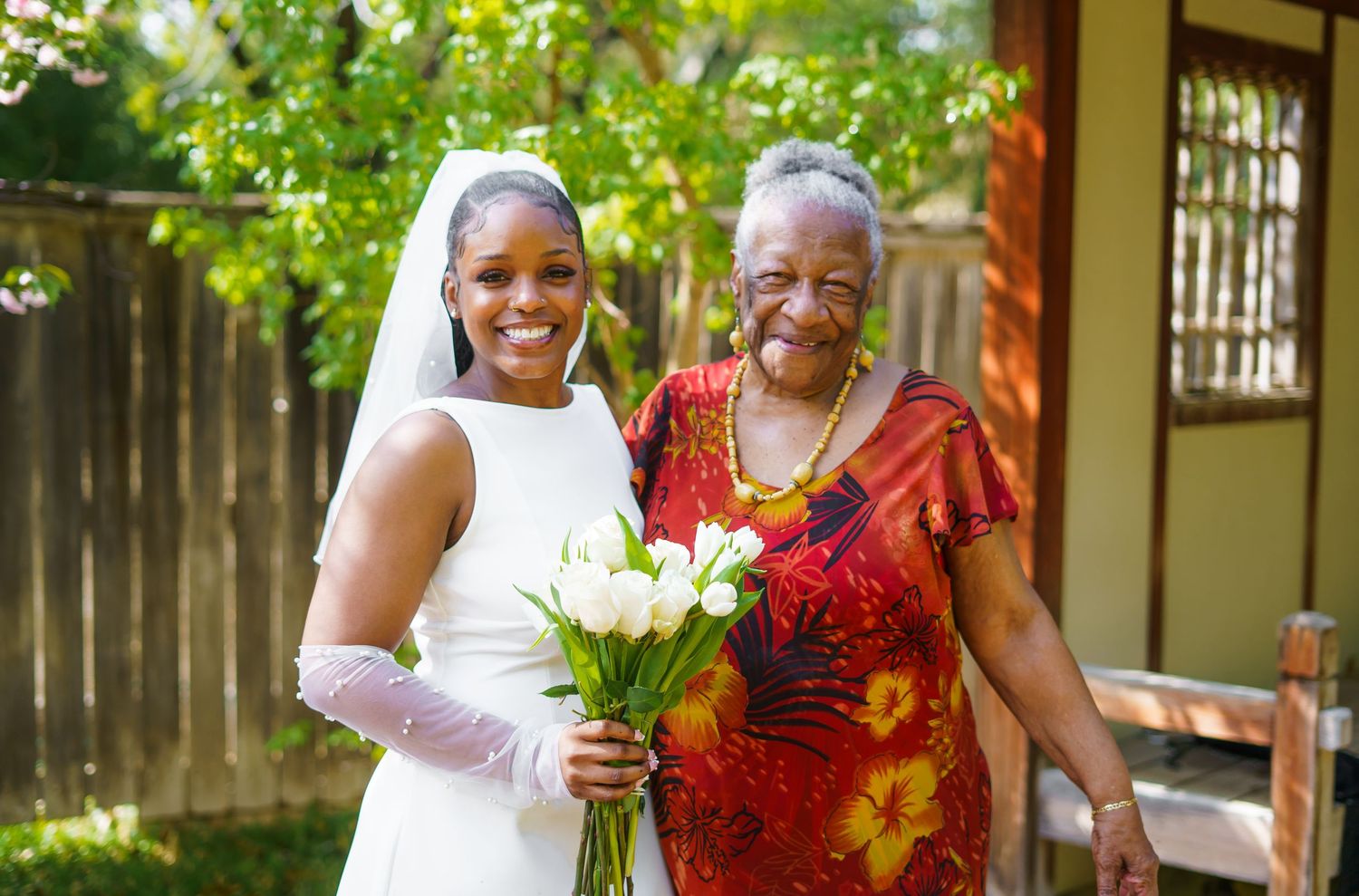 Smiling bride in white dress and veil holding tulip bouquet poses with elderly woman in red dress outdoors.