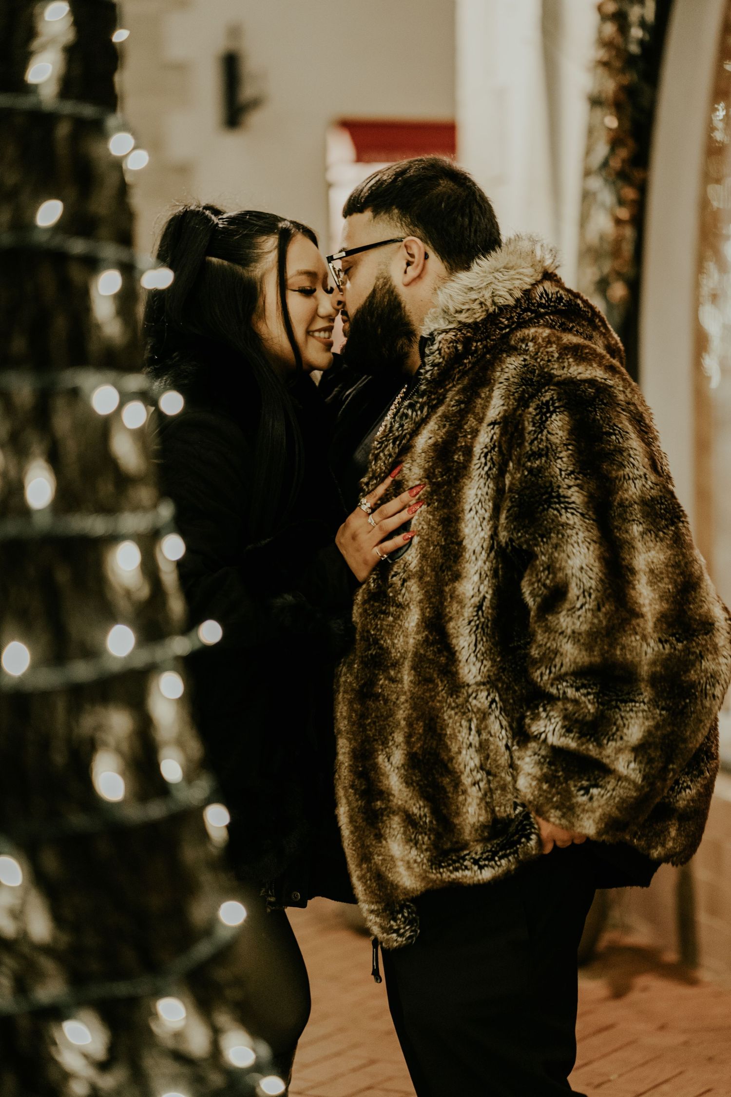 Couple sharing a tender kiss near a spiral light-wrapped tree on a festive brick walkway at night.