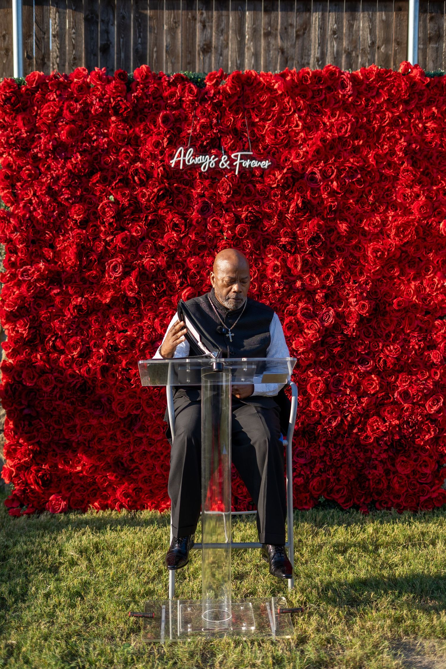 Wedding officiant speaks at clear podium before stunning red rose flower wall backdrop reading Always & Forever.