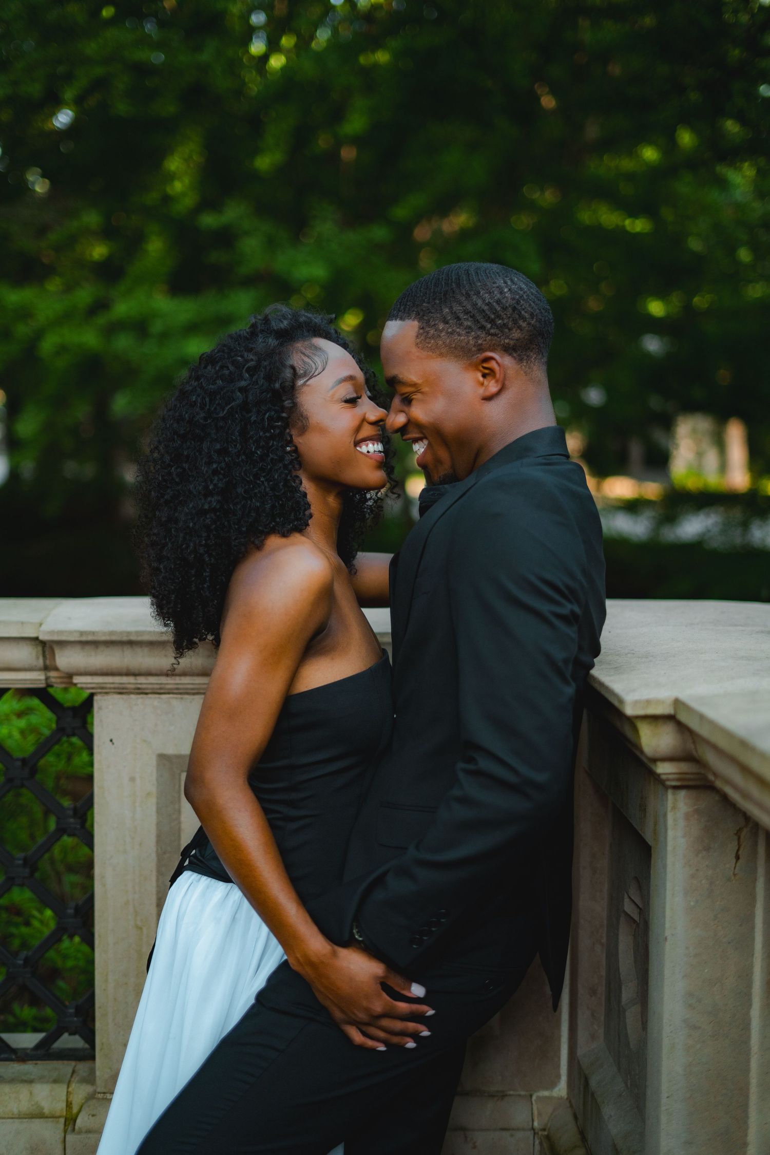 Joyful Black couple laughing and embracing by stone railing in elegant formal attire.