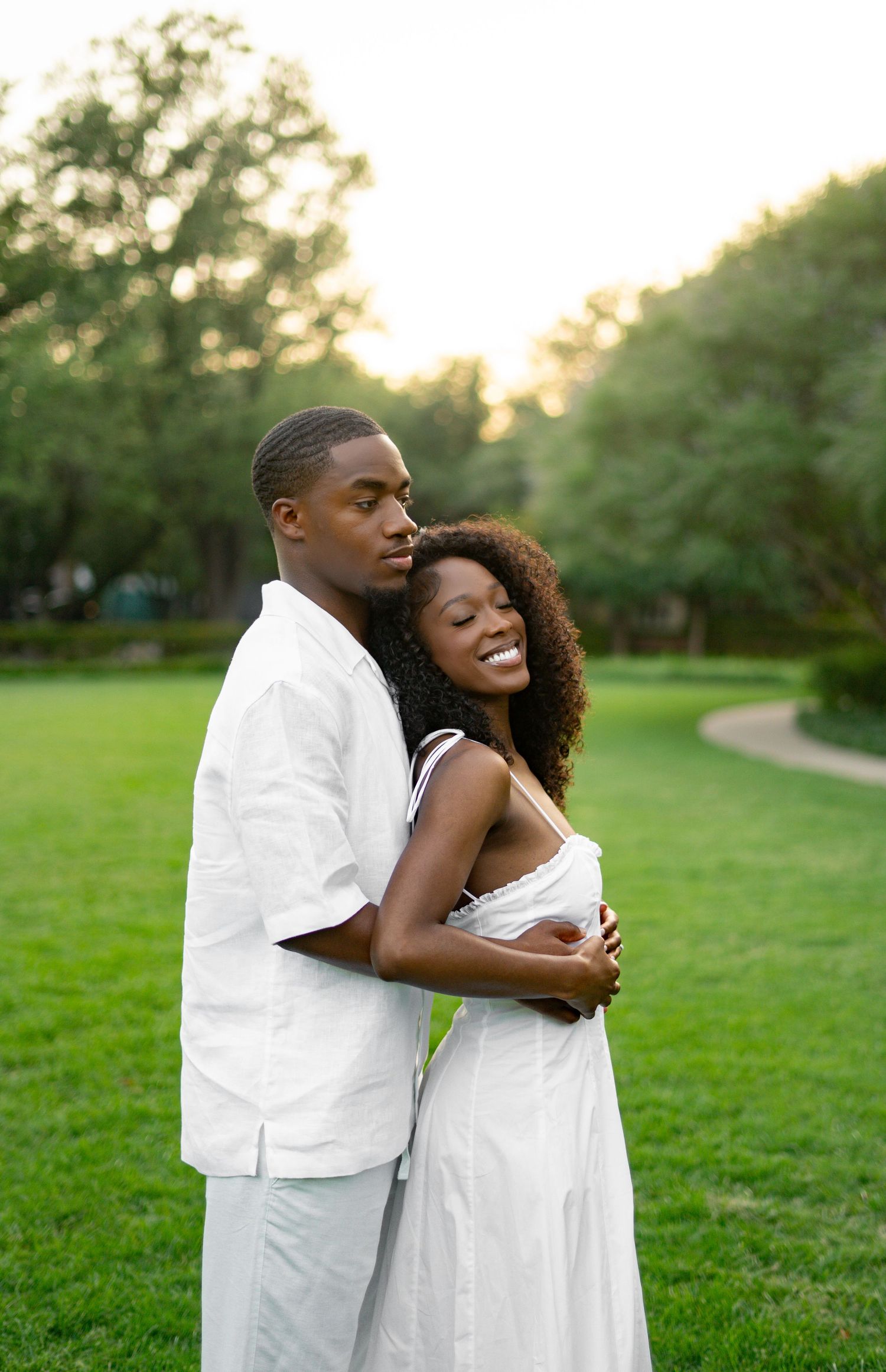 Couple in matching white outfits embracing on a lush green lawn at golden hour sunset.