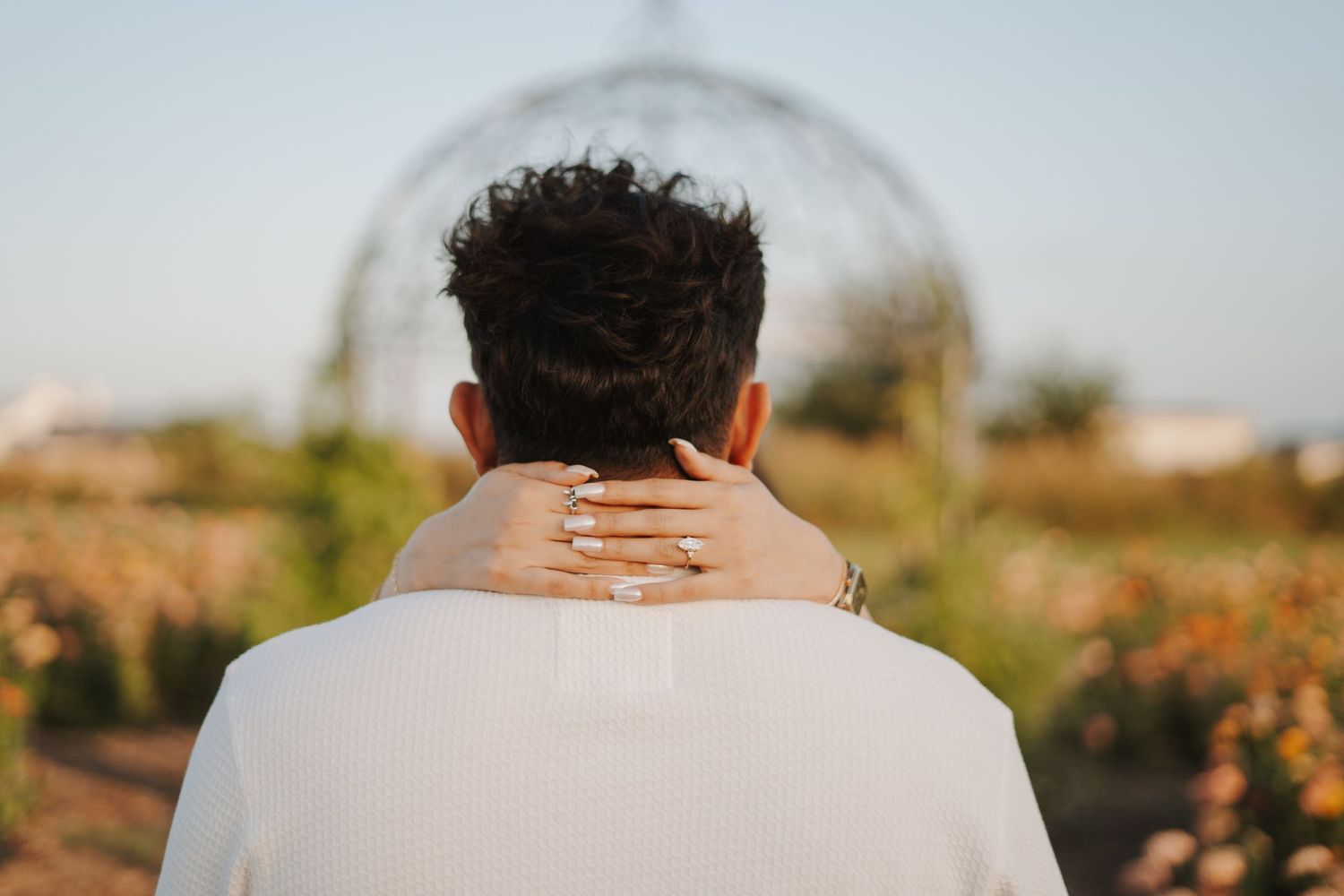 Couple embracing outdoors, woman's hand with ring on man's neck, garden arch background at golden hour.