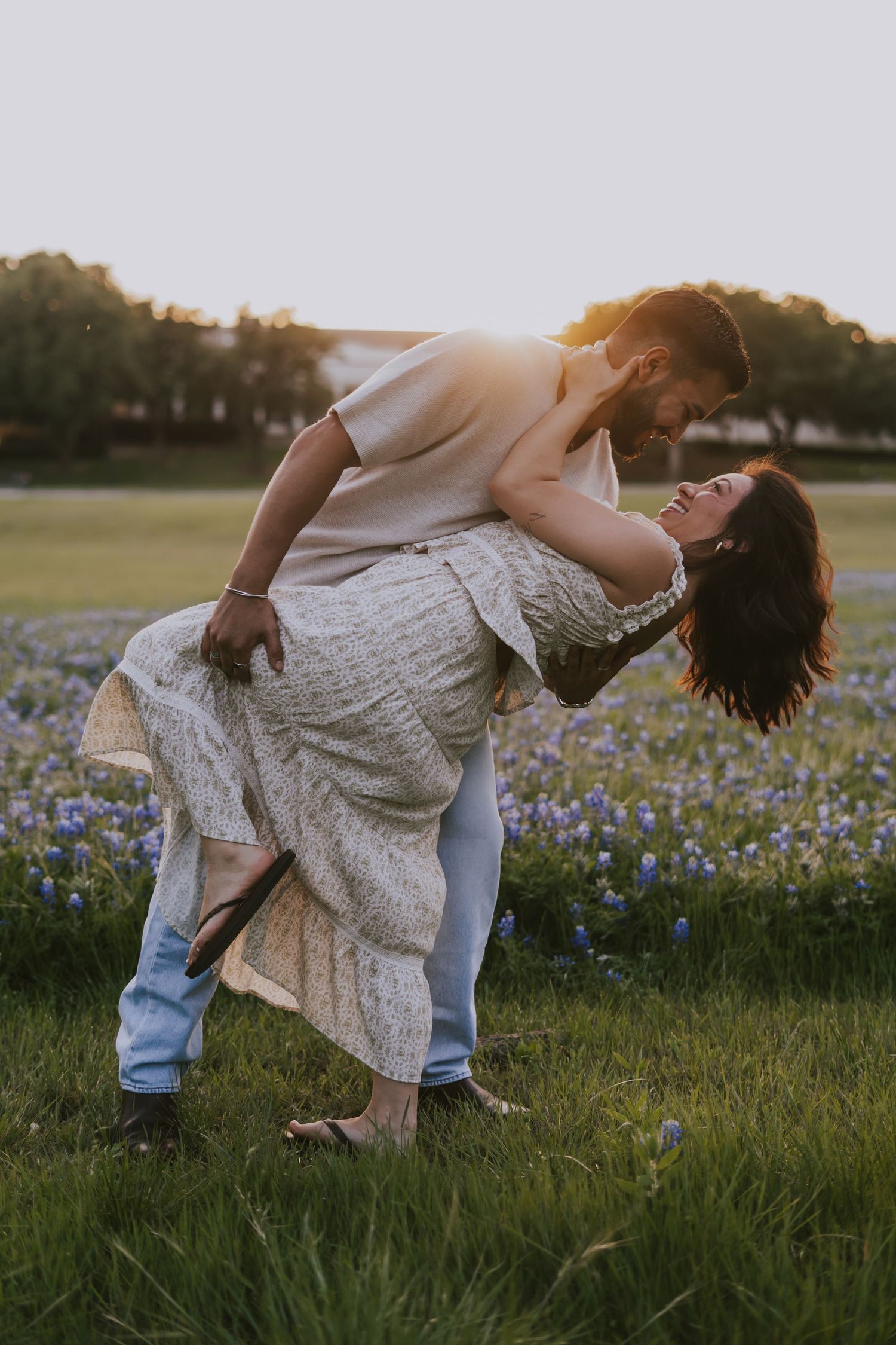 Couple dancing romantically in a blooming bluebonnet field at golden hour sunset, man dipping woman in floral dress.