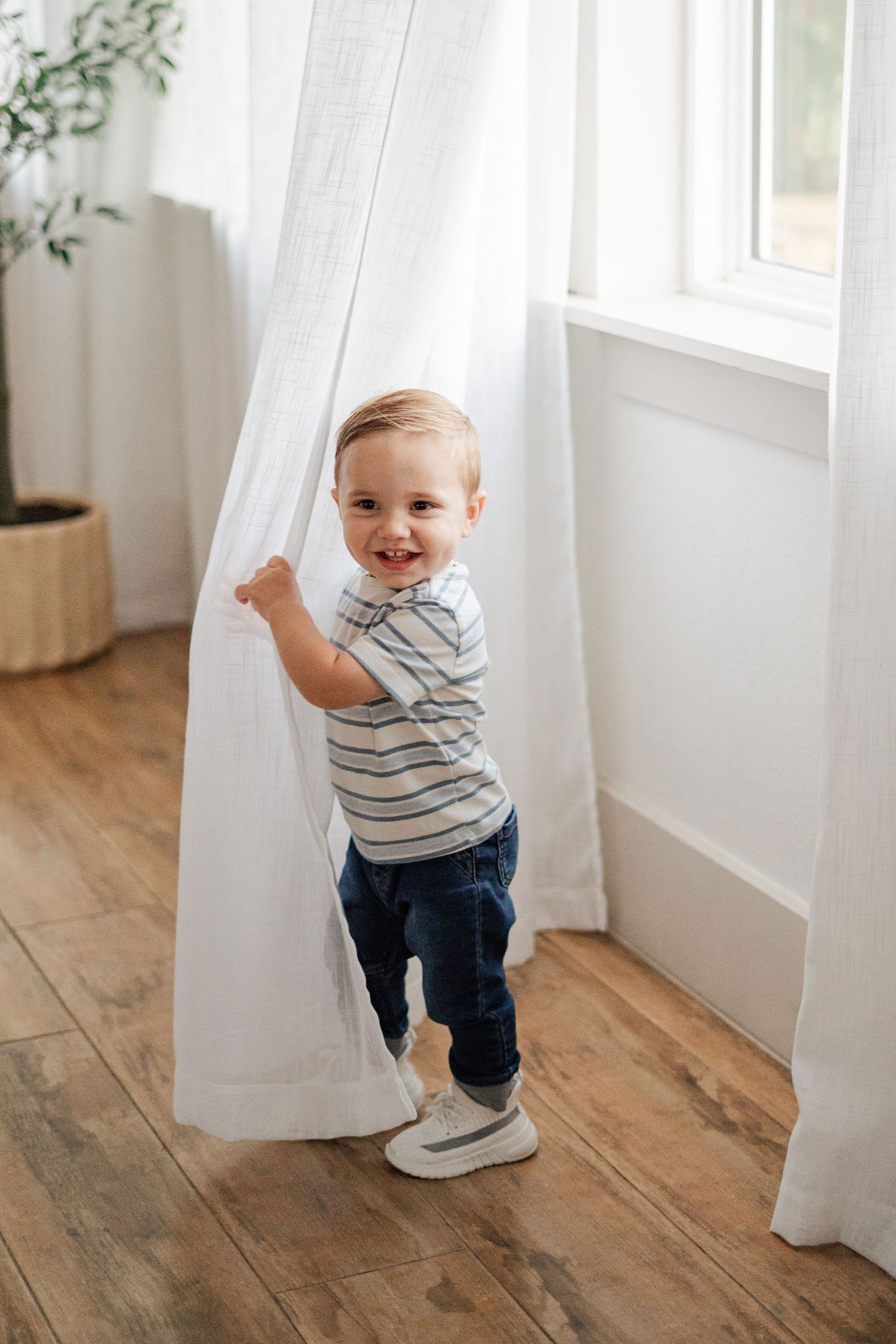 Smiling toddler in striped shirt and jeans playfully holding a white curtain near a bright window.