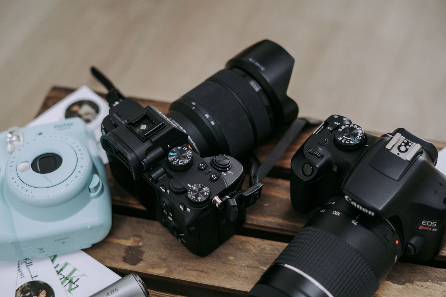 Three cameras on a wooden surface including a Fujifilm Instax Mini, mirrorless camera, and Canon DSLR.