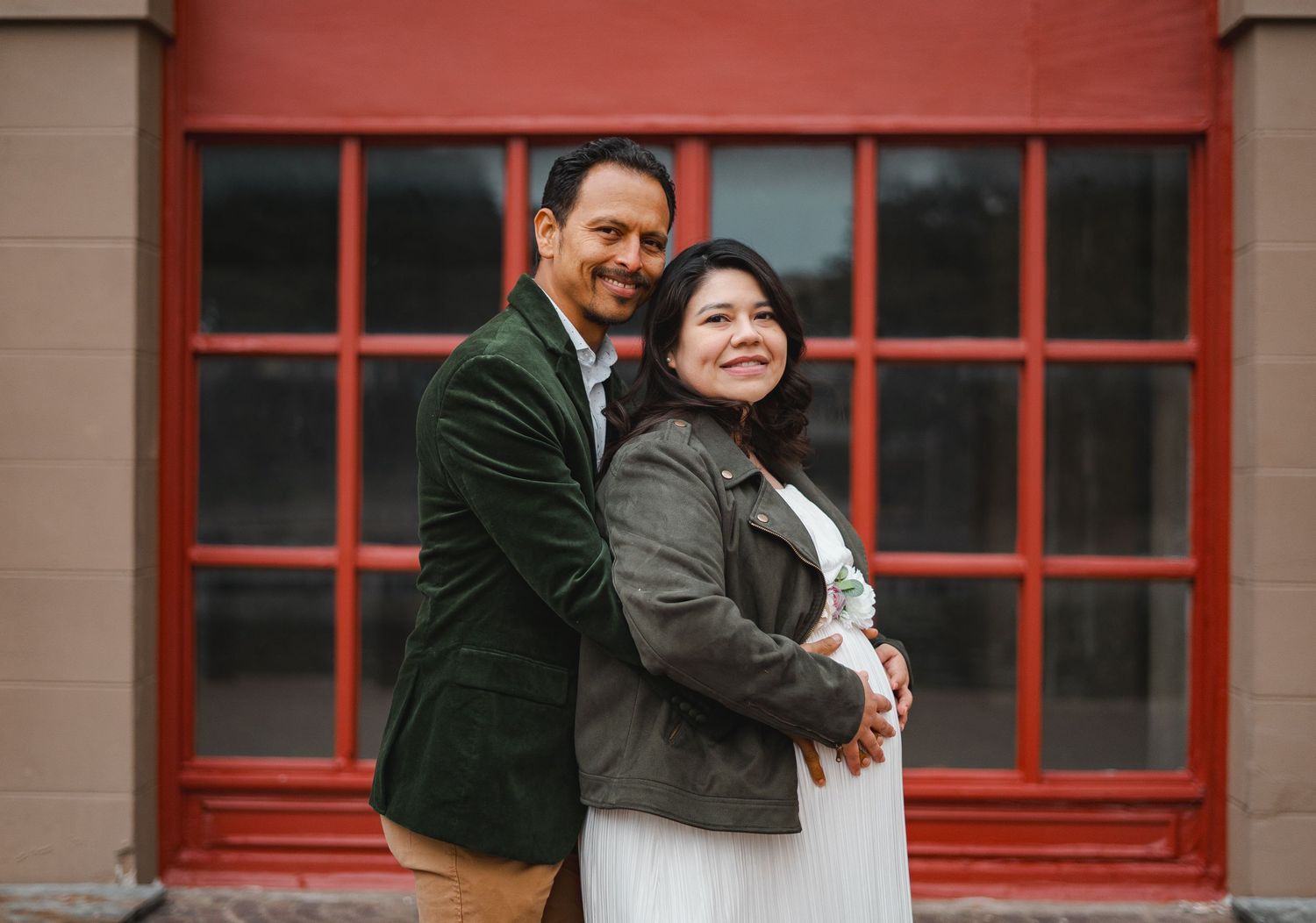 Couple embracing during maternity photo session in front of vibrant red building exterior.