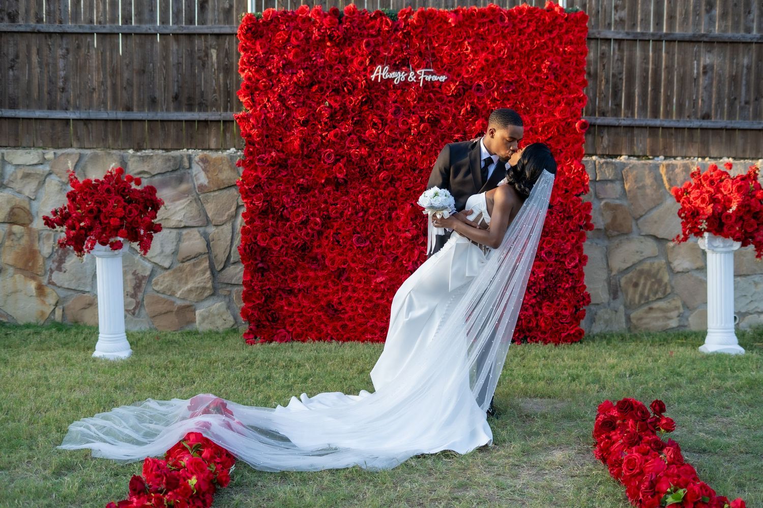 Groom dips bride in romantic pose before red rose flower wall with white floral arrangements.