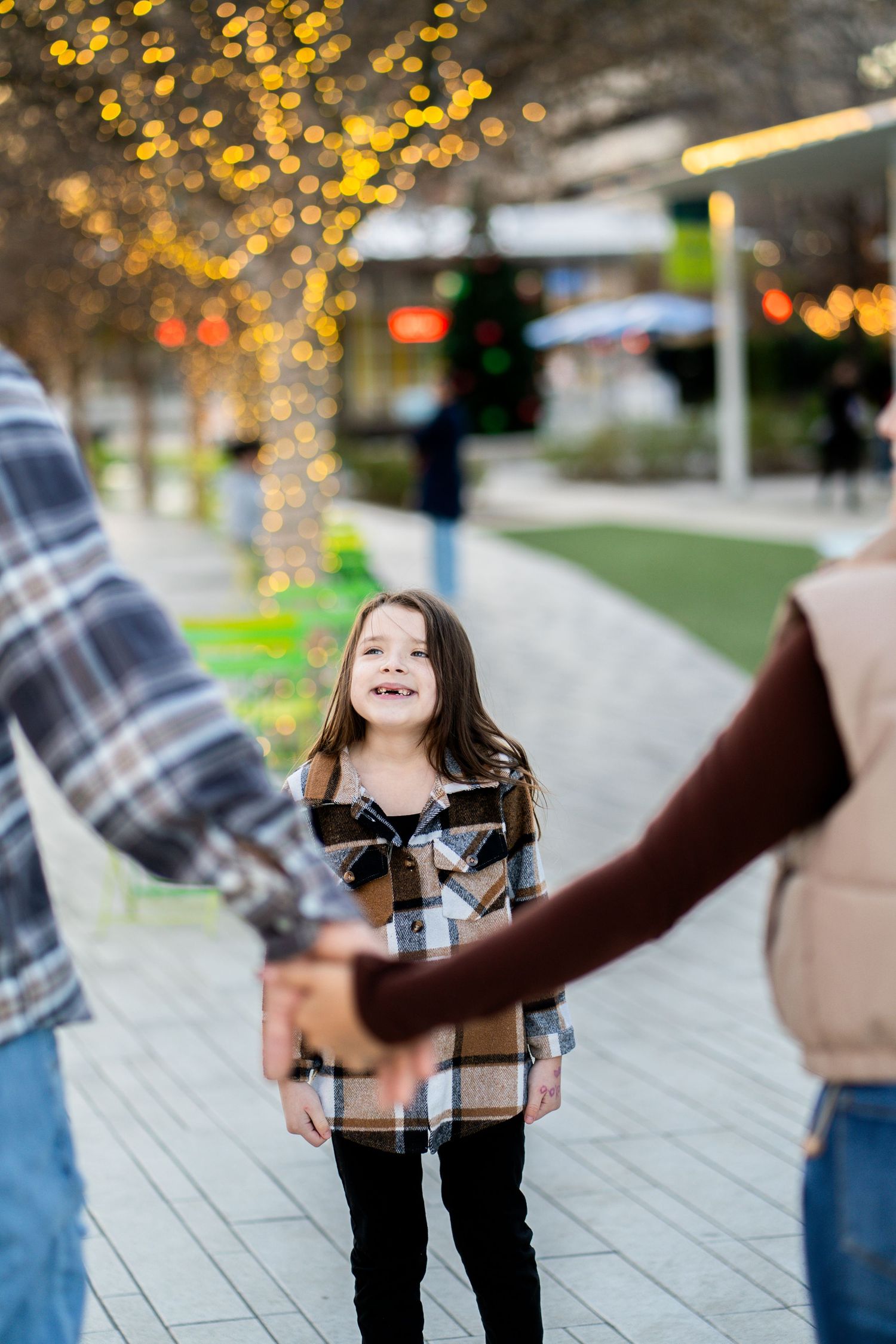 A young girl looks up joyfully while holding hands with her parents on a brick pathway.