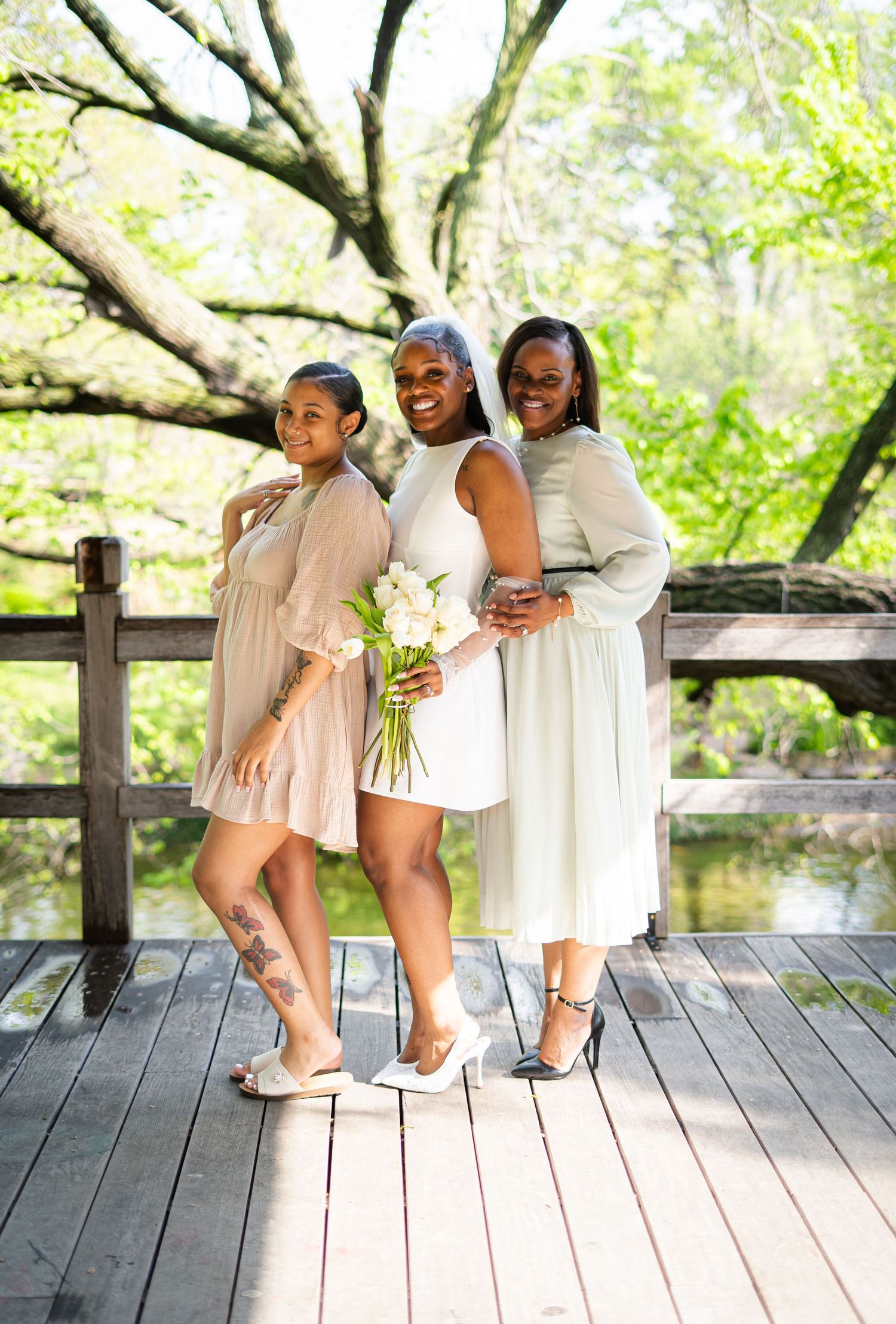Bride in white mini dress and veil poses with two women on a wooden deck surrounded by trees.