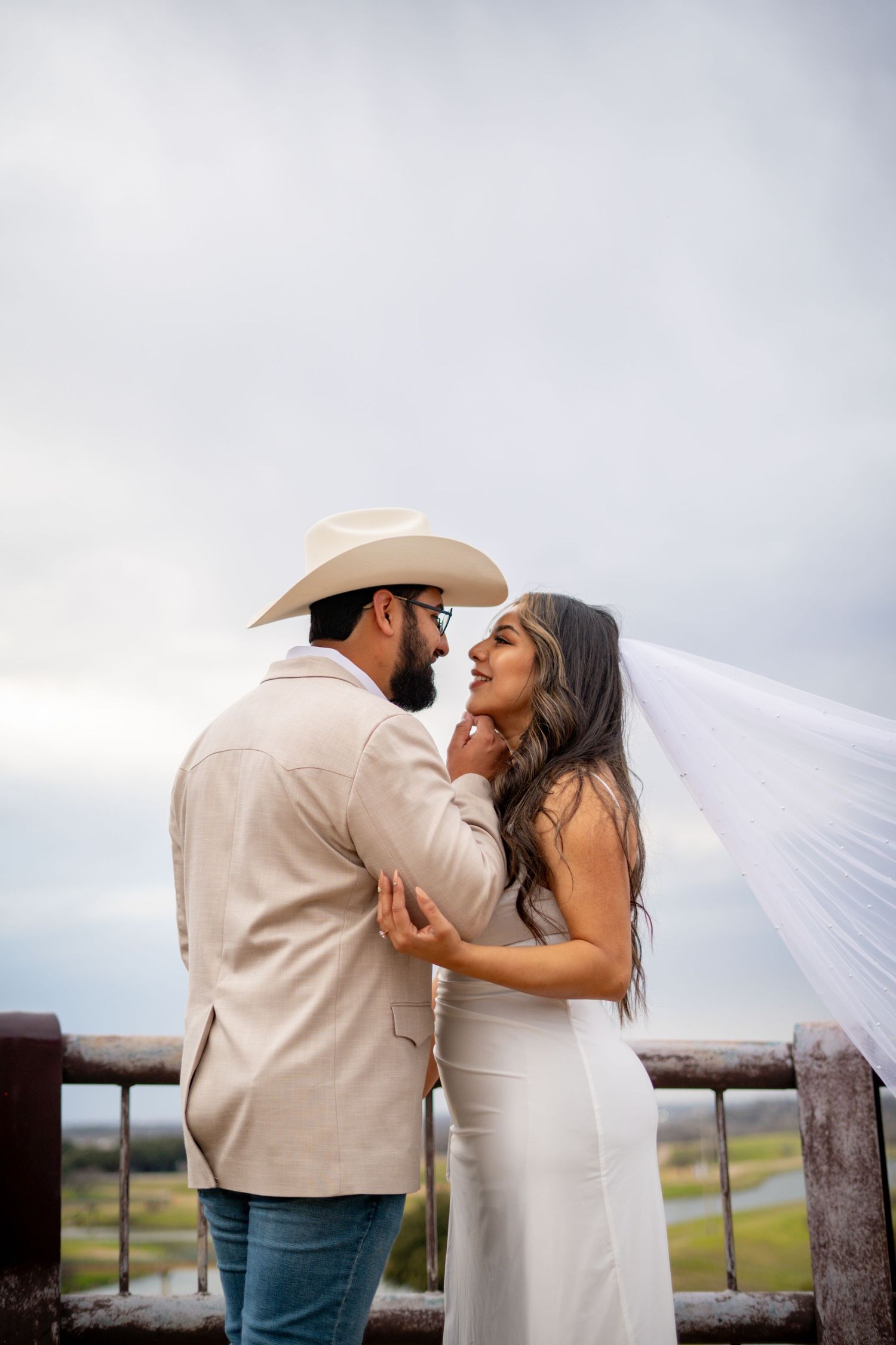 A bride in a white gown with flowing veil shares a romantic moment with her cowboy hat-wearing groom outdoors.