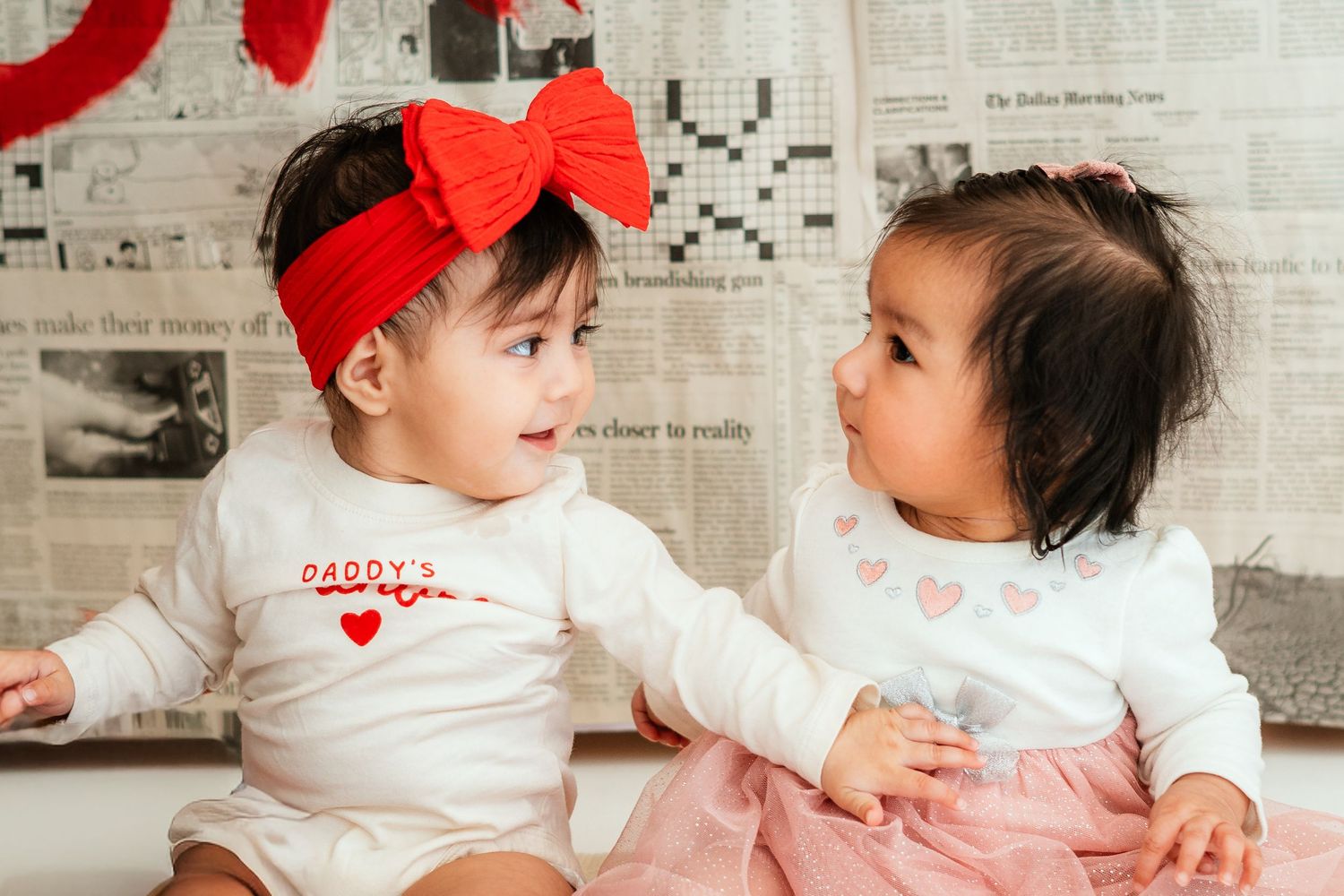 Two adorable baby girls in Valentine outfits smiling at each other indoors.