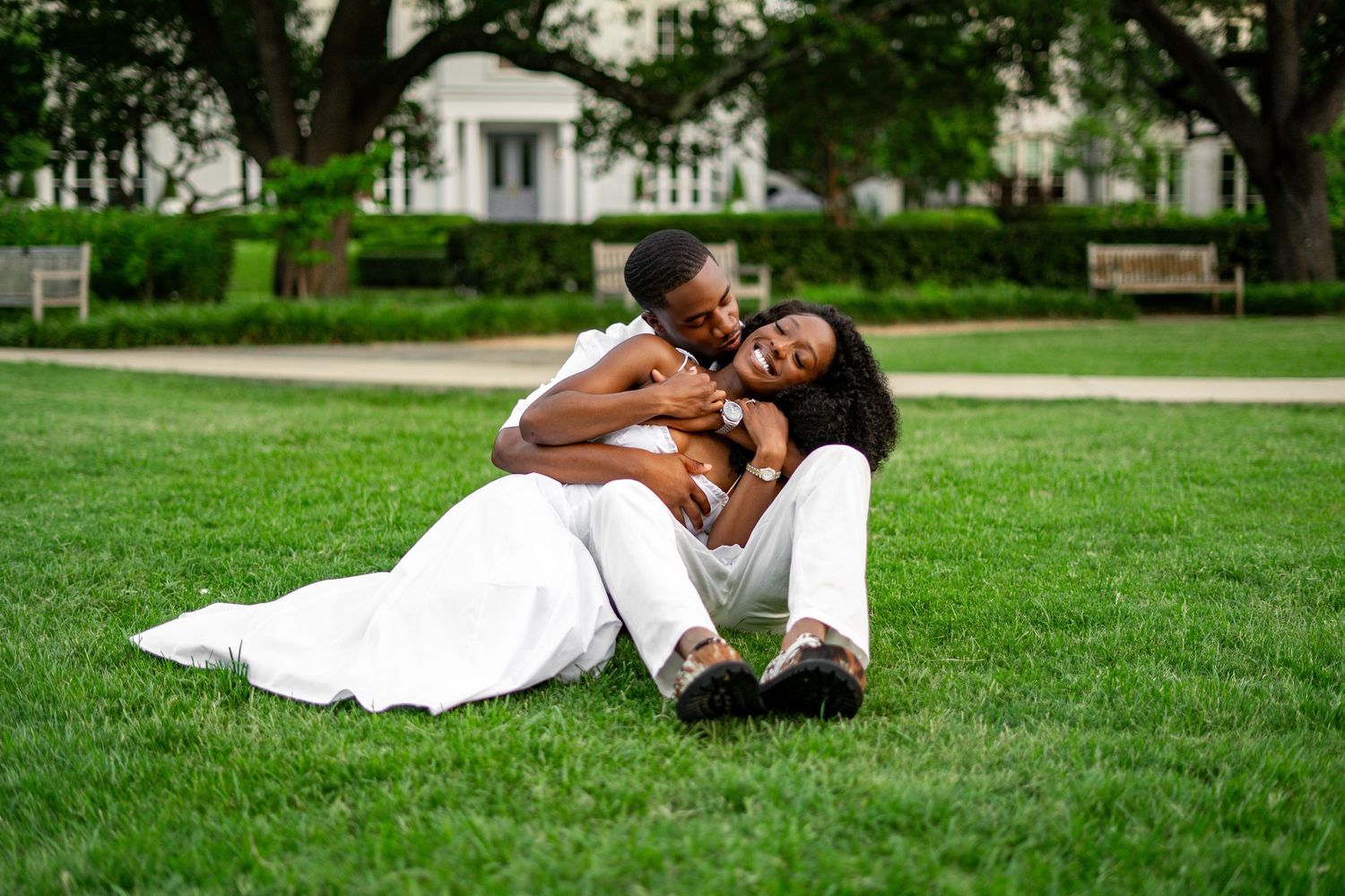 A joyful Black couple in white outfits playfully embrace on lush green grass during an outdoor engagement session.