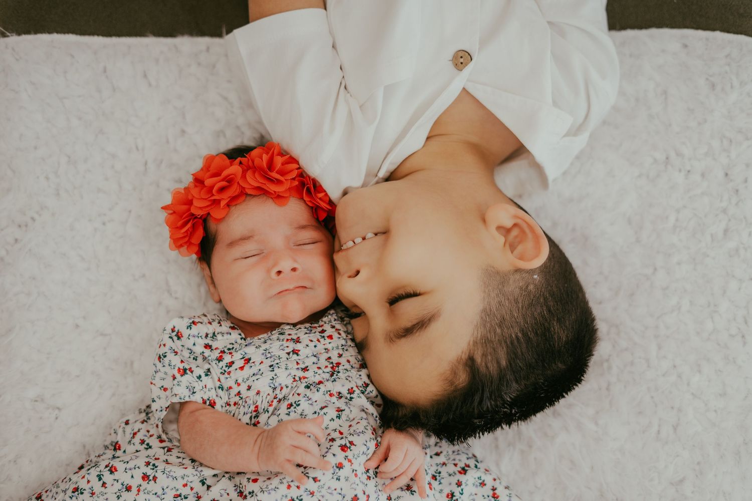 Older sibling gently kissing newborn baby girl wearing red floral headband.