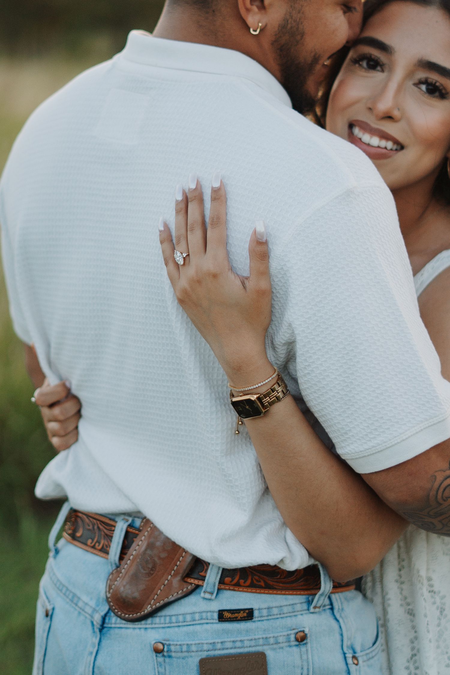Couple embracing outdoors, woman smiling showing engagement ring, wearing watch, man in white shirt and jeans with leather belt.