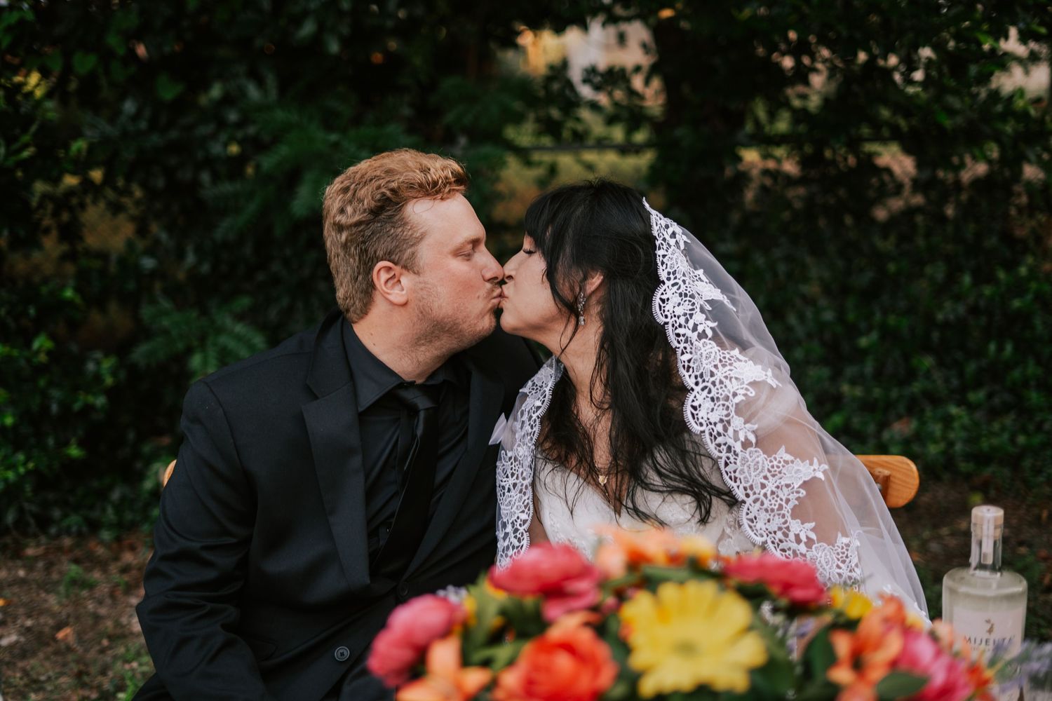 Newlywed couple shares first kiss at outdoor wedding reception surrounded by colorful floral arrangements.